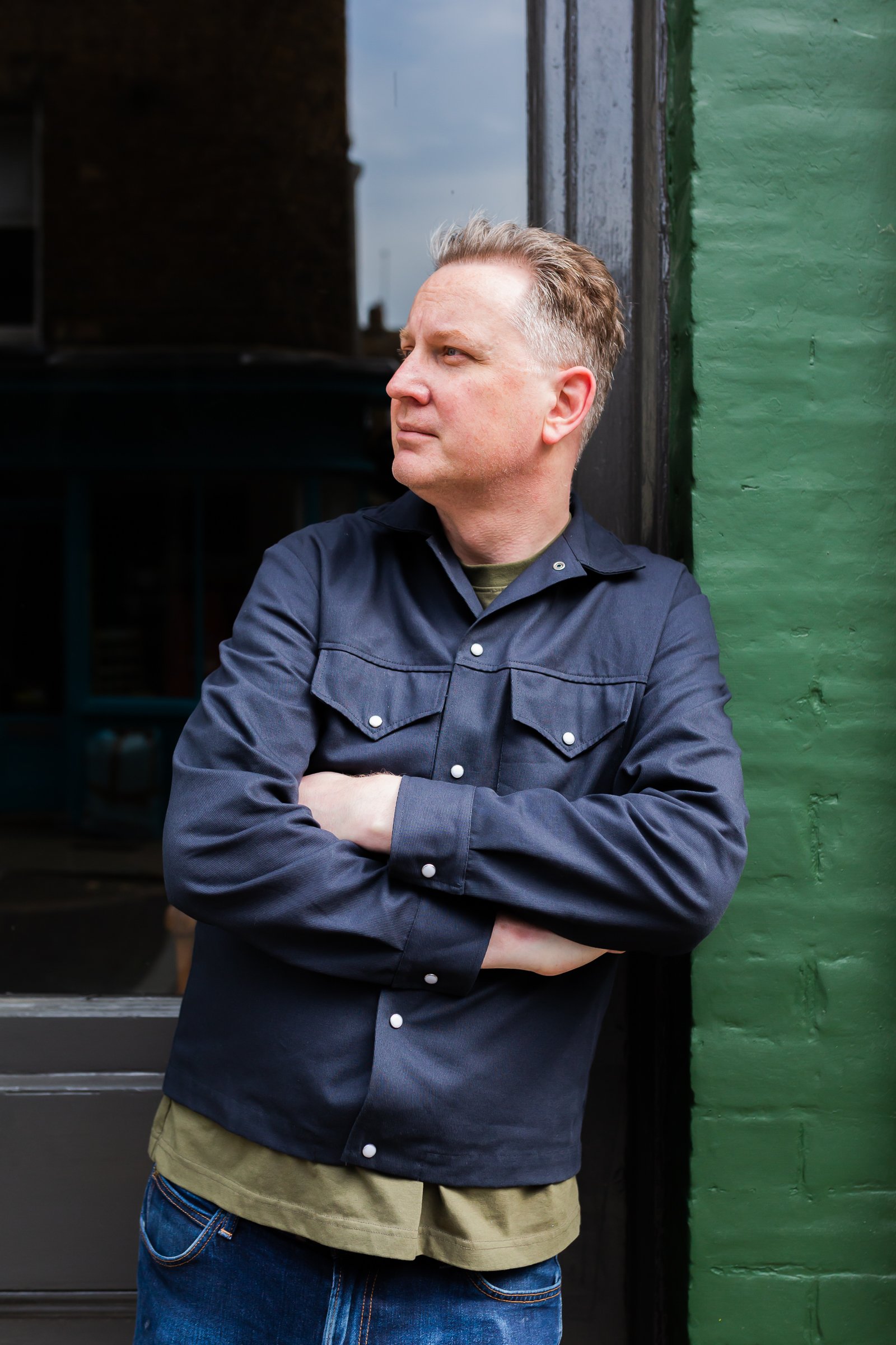 A man with short blond hair wearing a dark button-up shirt with rolled sleeves over a green T-shirt, standing against a green brick wall, arms crossed, looking to the left.