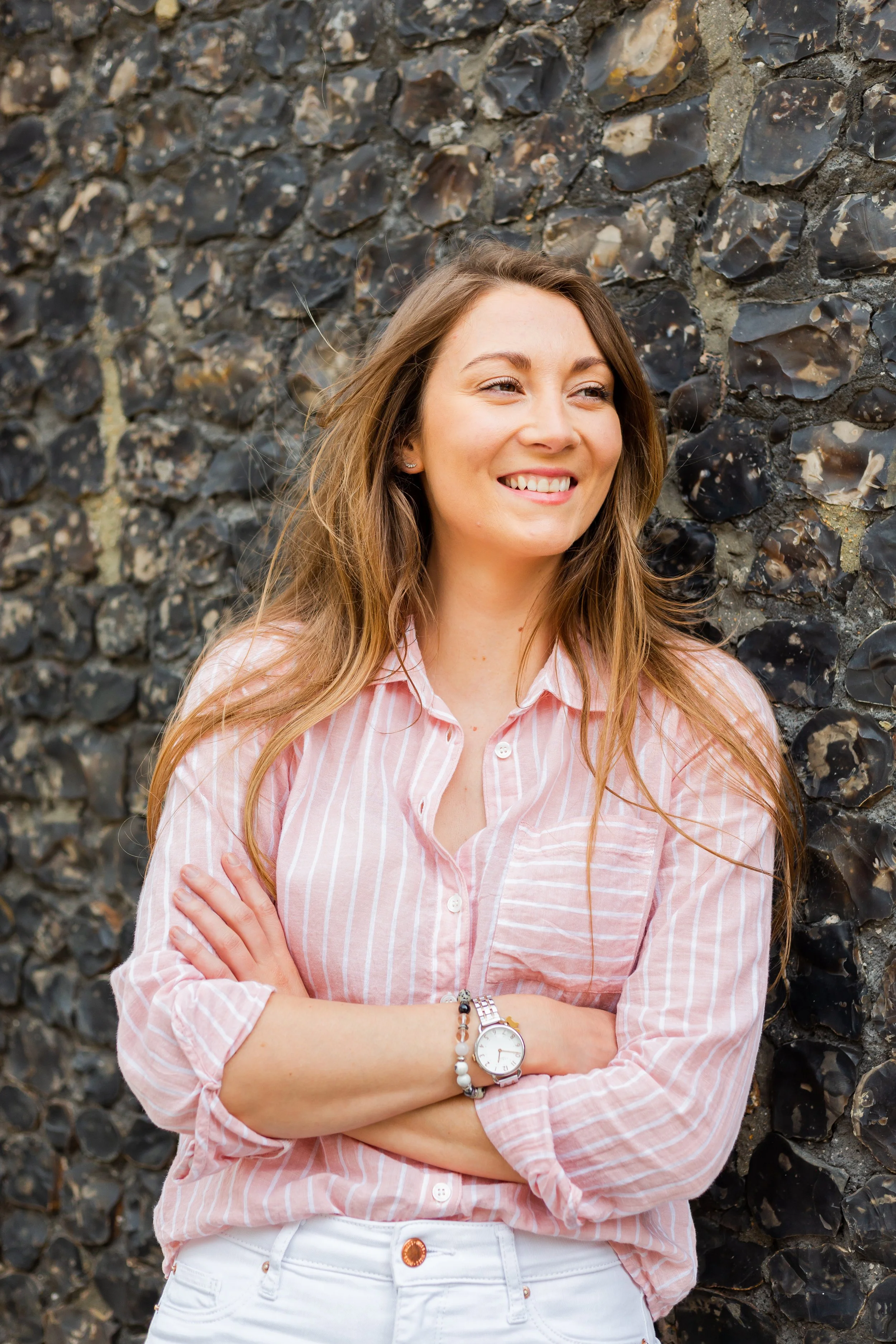 A young woman with long light brown hair is smiling and standing with arms crossed in front of a textured stone wall, wearing a pink and white striped button-down shirt, white pants, and a wristwatch.