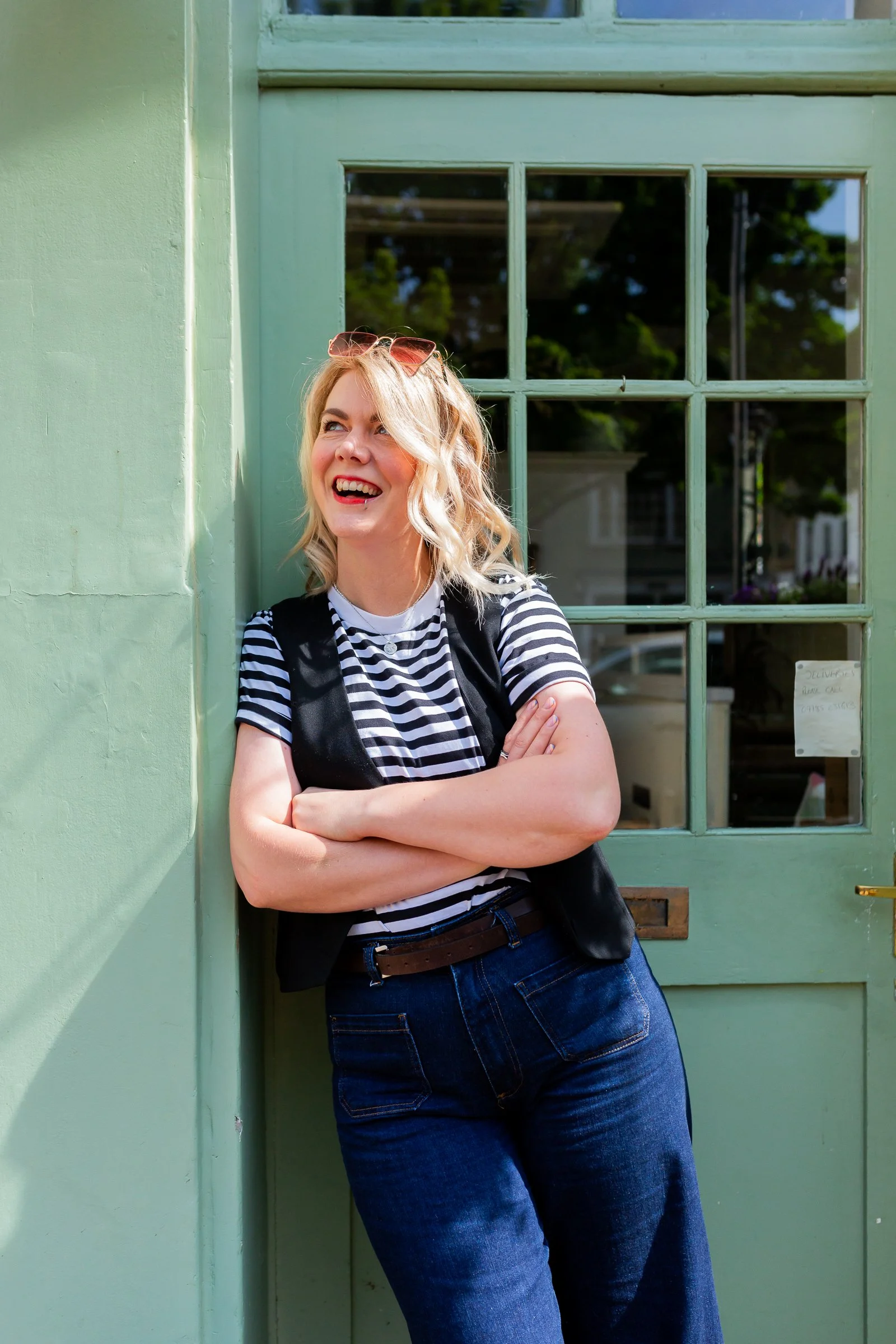 Girl with blonde curly hair and sunglasses smiling, standing with arms crossed outside a green door.