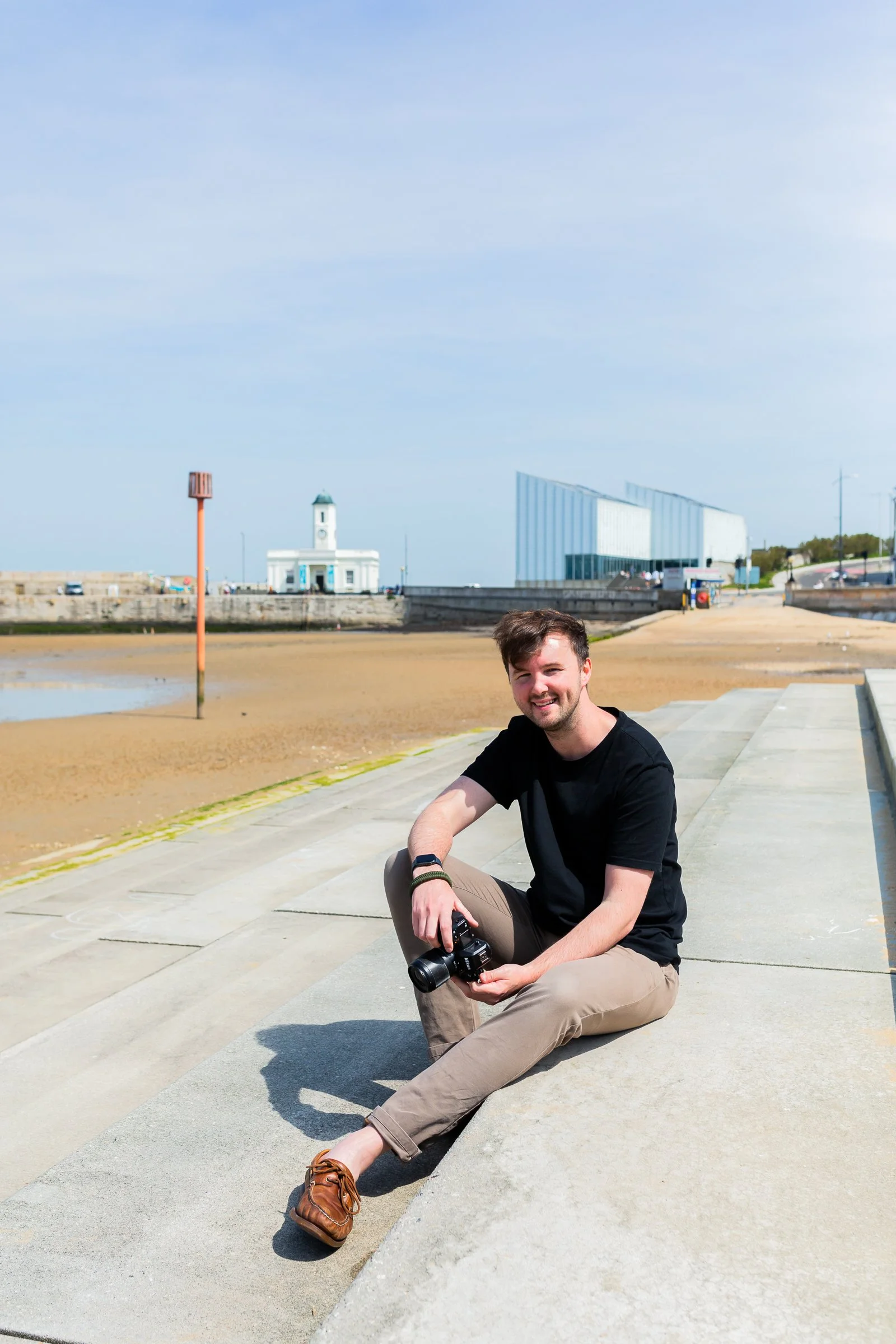 A young man with brown hair, wearing a black T-shirt, tan pants, and brown shoes, sitting on concrete steps near a beach, holding a camera and smiling.