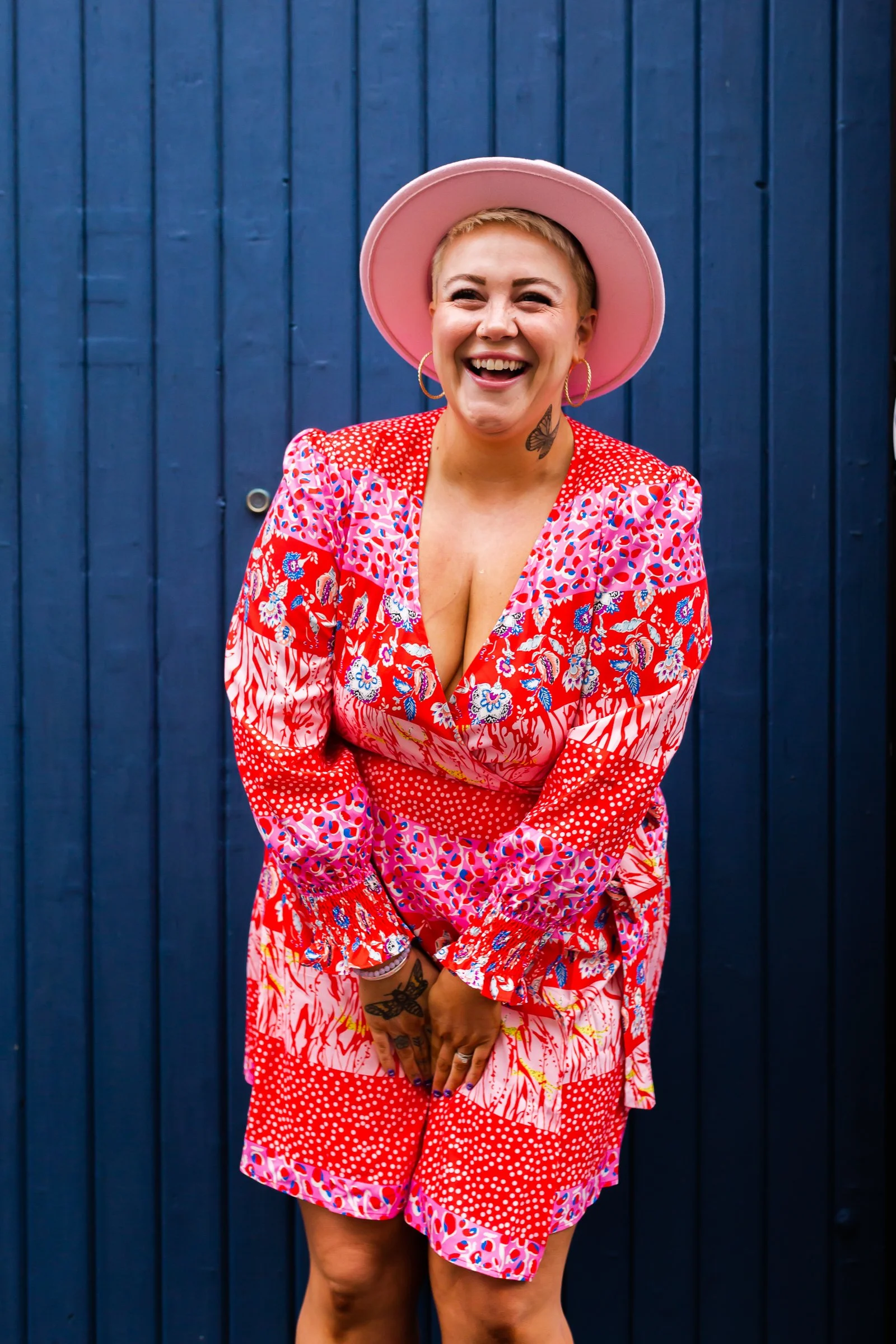 A woman laughing, wearing a pink hat and a red, pink, and blue patterned dress, standing in front of a dark blue wooden wall.