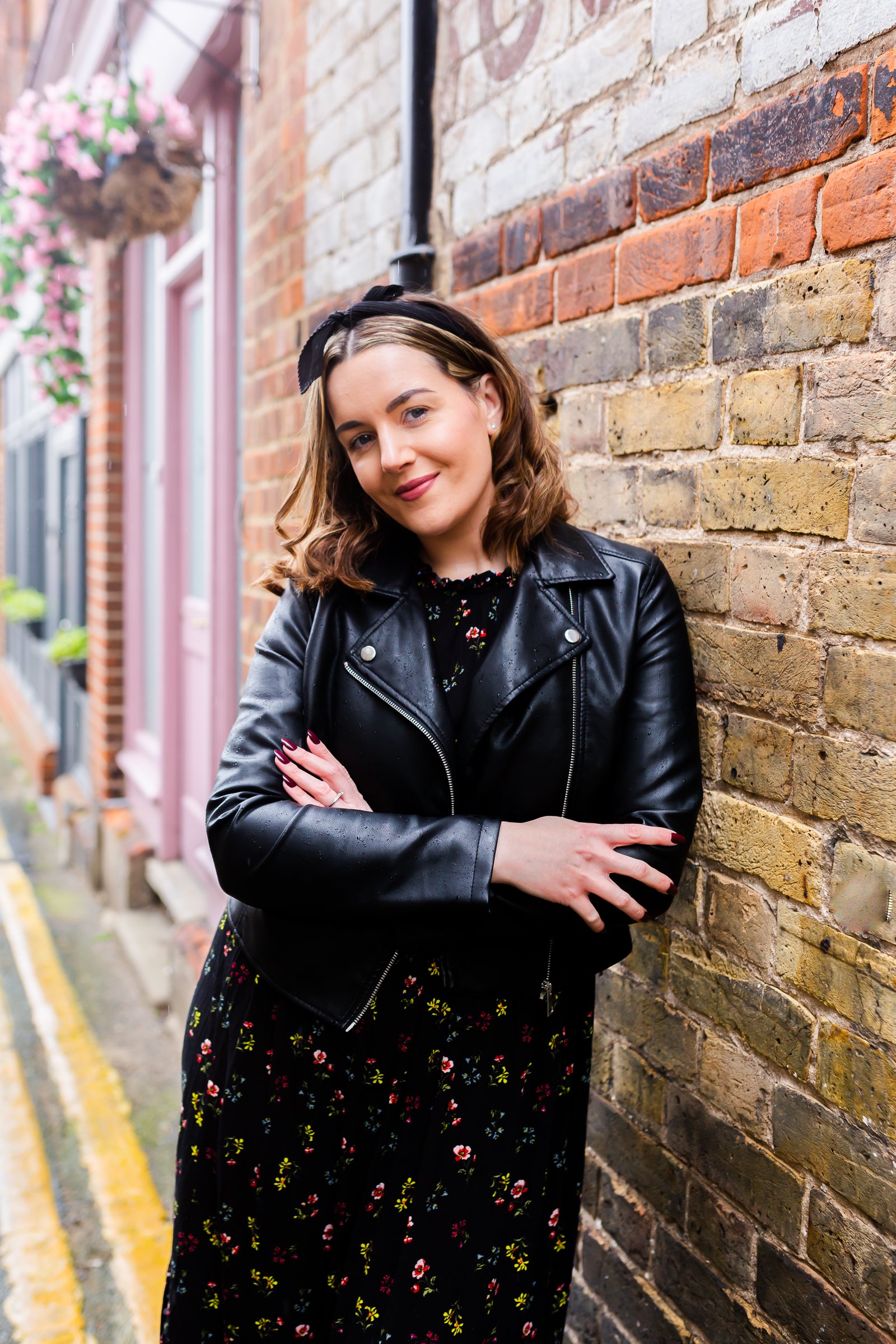 A woman with shoulder-length brown hair wearing a black leather jacket, floral dress, and black headband standing against a brick wall with her arms crossed and smiling.