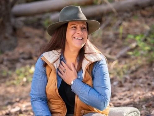 A woman outdoors wearing a gray hat, a blue denim jacket, and a tan vest, smiling with her hand on her chest.