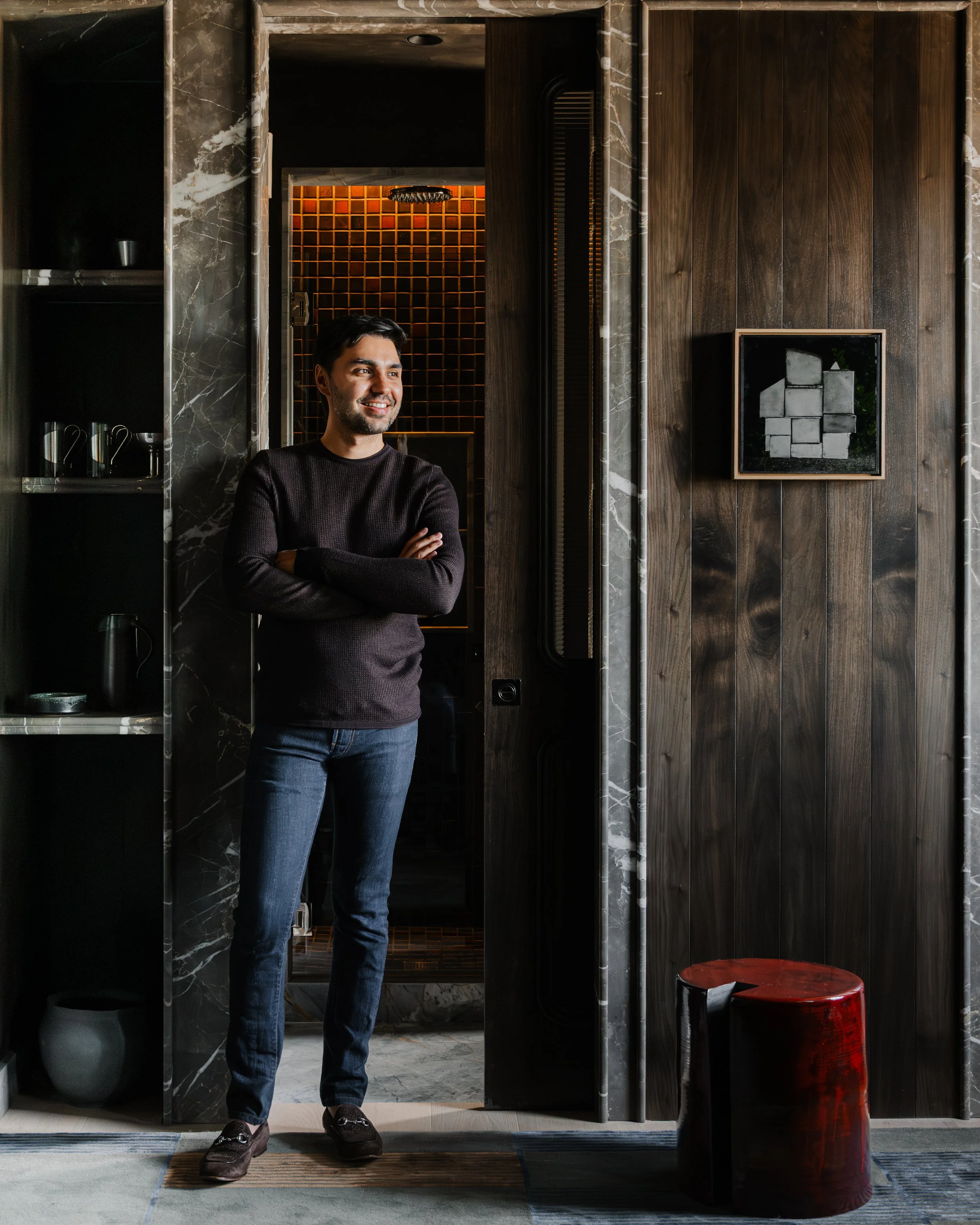 A man sitting on a wooden bench in a modern, dark-colored kitchen, with a vase of purple flowers and green apples on the countertop behind him.