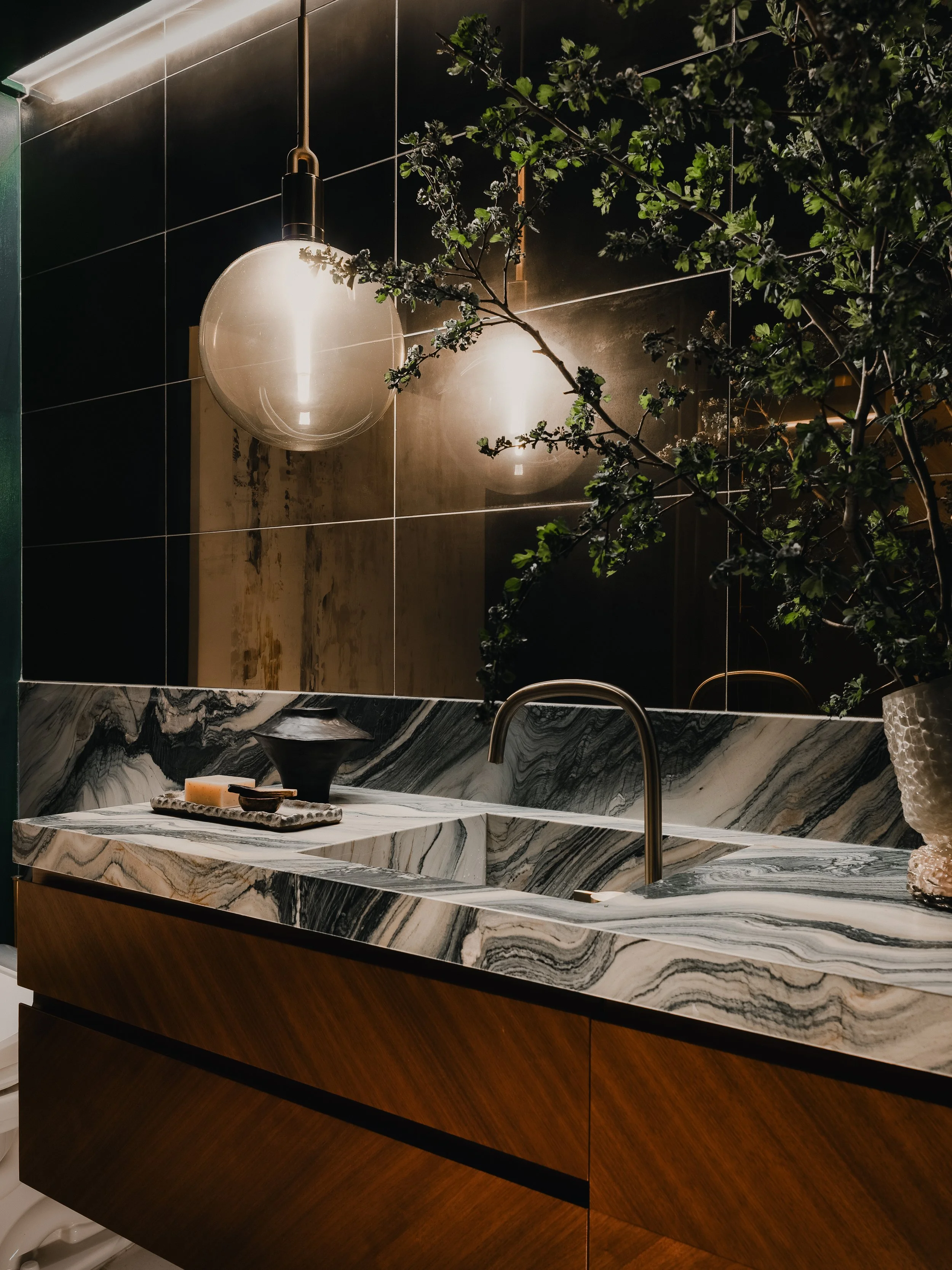 A minimalist bathroom with a toilet, a wooden vanity with a marble top, a mirror, with two wall-mounted globe lights on either side of the mirror. There is a towel hanging on a bar to the left and a wooden door to the right.