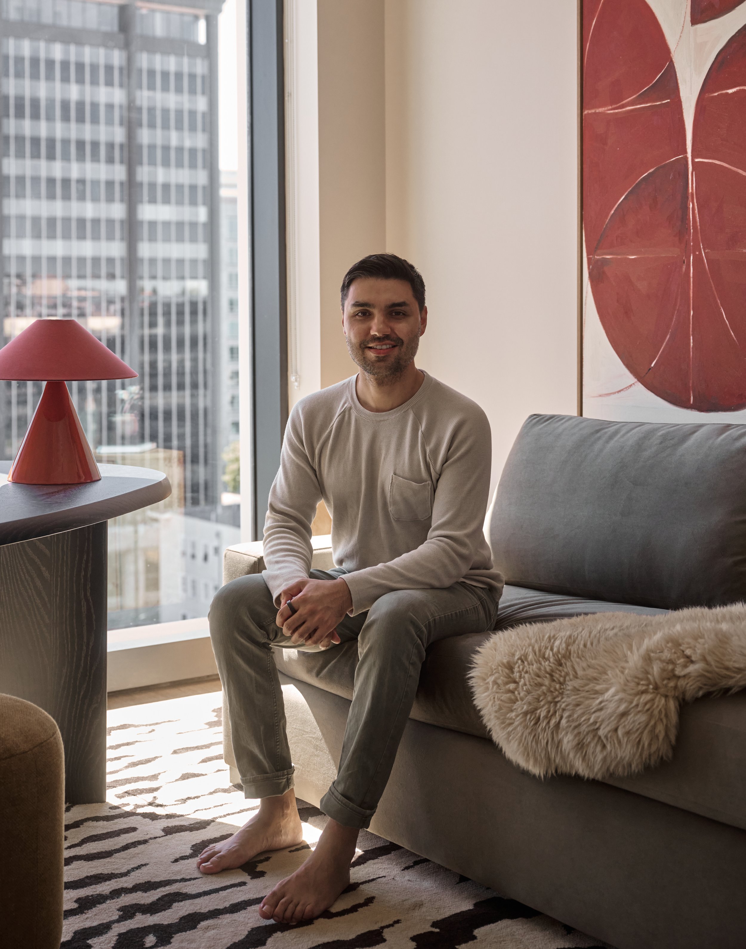 A man sitting on a wooden bench in a modern, dark-colored kitchen, with a vase of purple flowers and green apples on the countertop behind him.