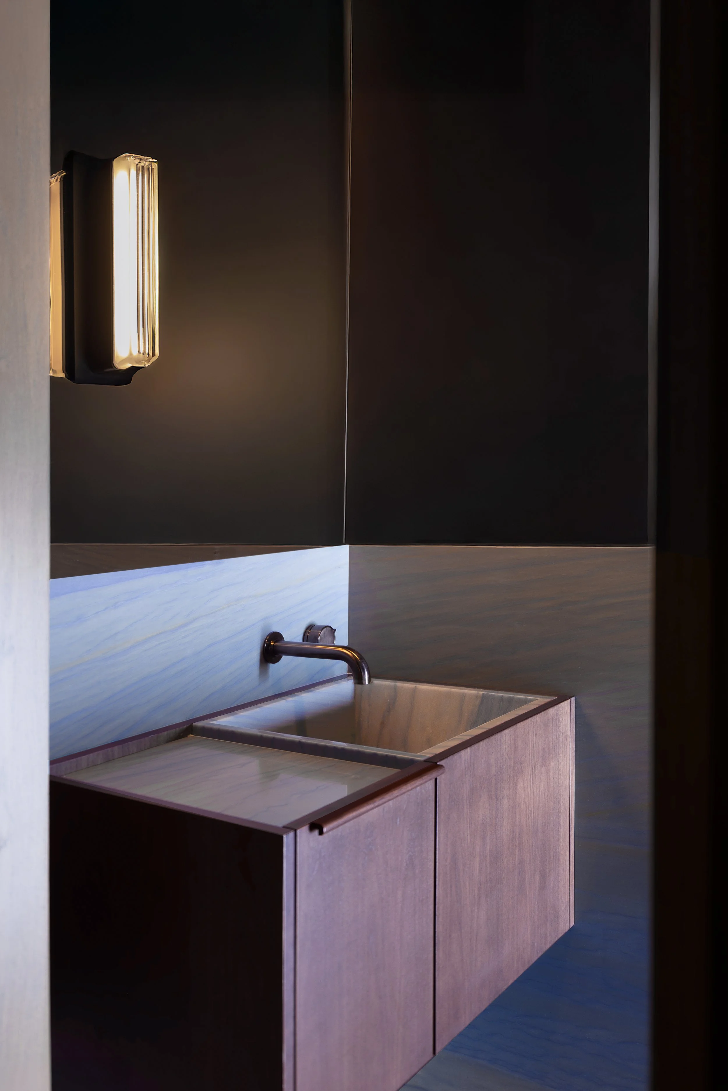 Modern laundry sink with wooden cabinet, black wall, and wall-mounted light fixture.