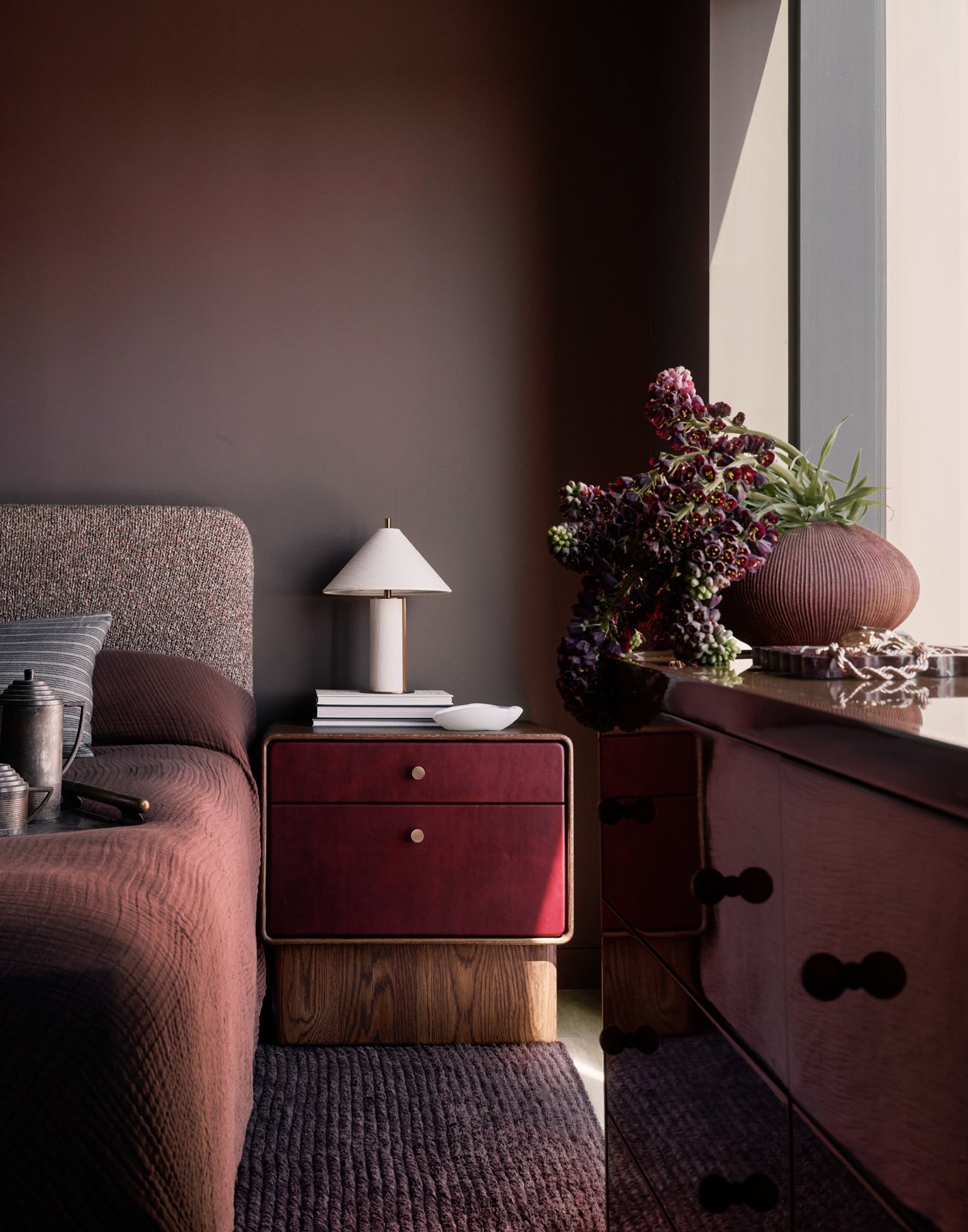 A modern wine bar with wooden cabinets, marble countertops, and shelves filled with wine bottles. Natural light from two large windows brightens the space, which features a small sink and decorative lighting.