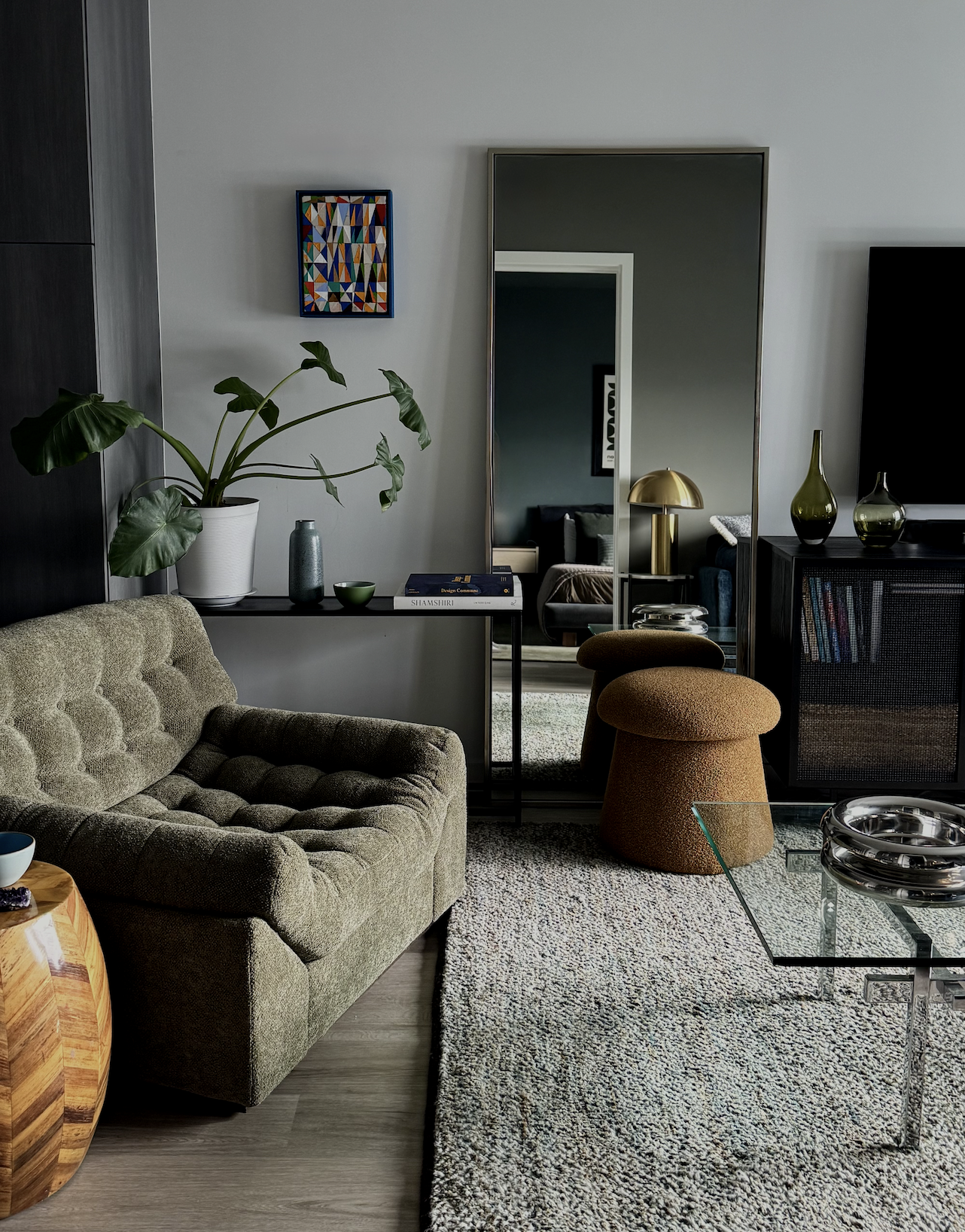 A modern living room with a beige velvet sofa, a large green potted plant, a tall mirror reflecting a doorway, a small black table with books, a gold desk lamp, and decorative vases on a black cabinet. A glass coffee table with decorative items sits over a textured rug.