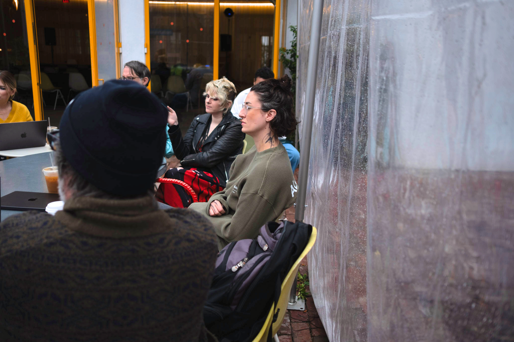  A group of people sitting outdoors at a table, attentively listening. They appear relaxed and engaged, with coffee cups and laptops on the table. Overcast lighting. 
