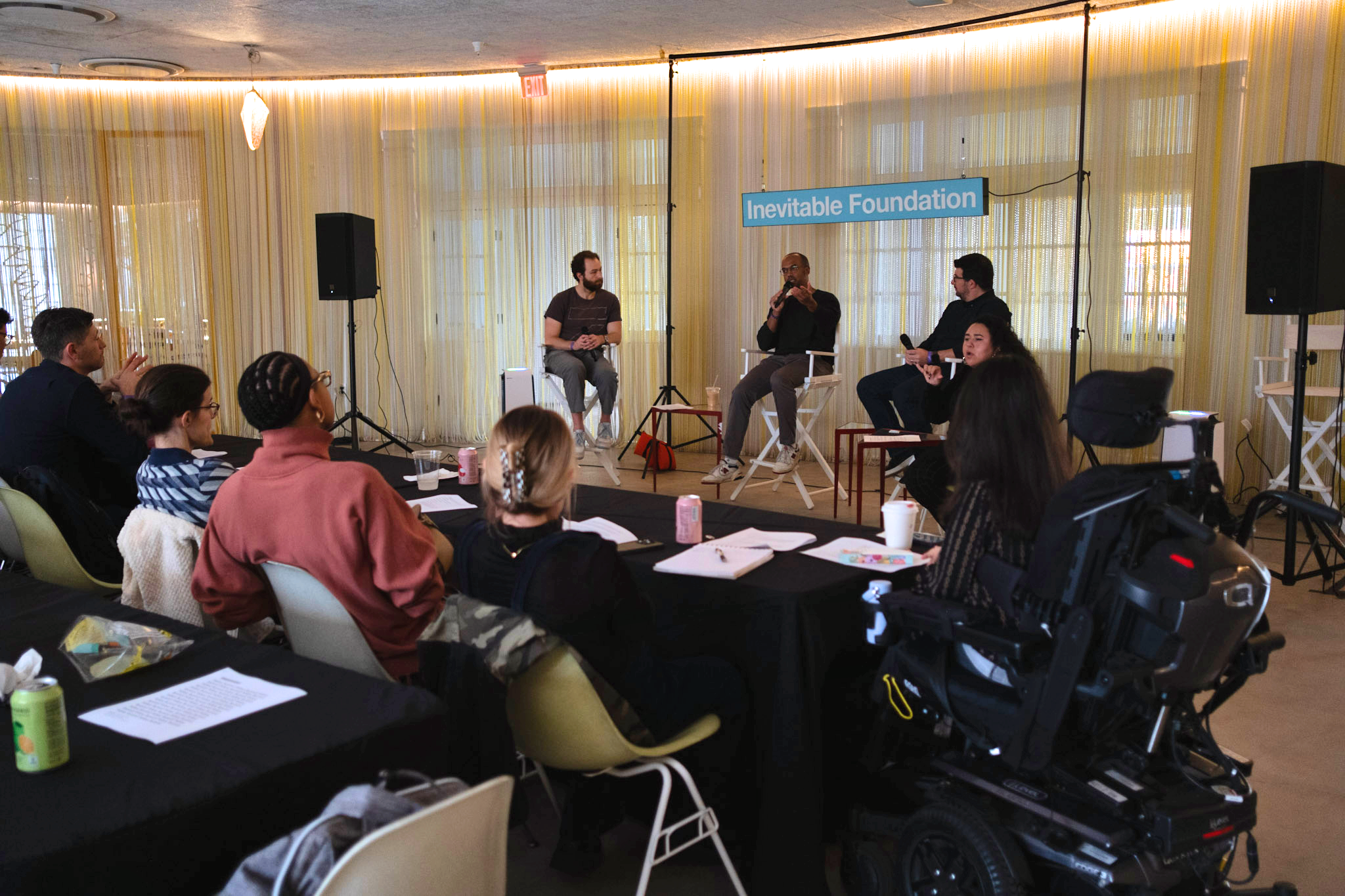  Three people sit in director’s chairs in front of an "Inevitable Foundation" sign as part of a panel discussion. In the foreground is a group of six diverse people, including one in a wheelchair, listening to the panelists. 