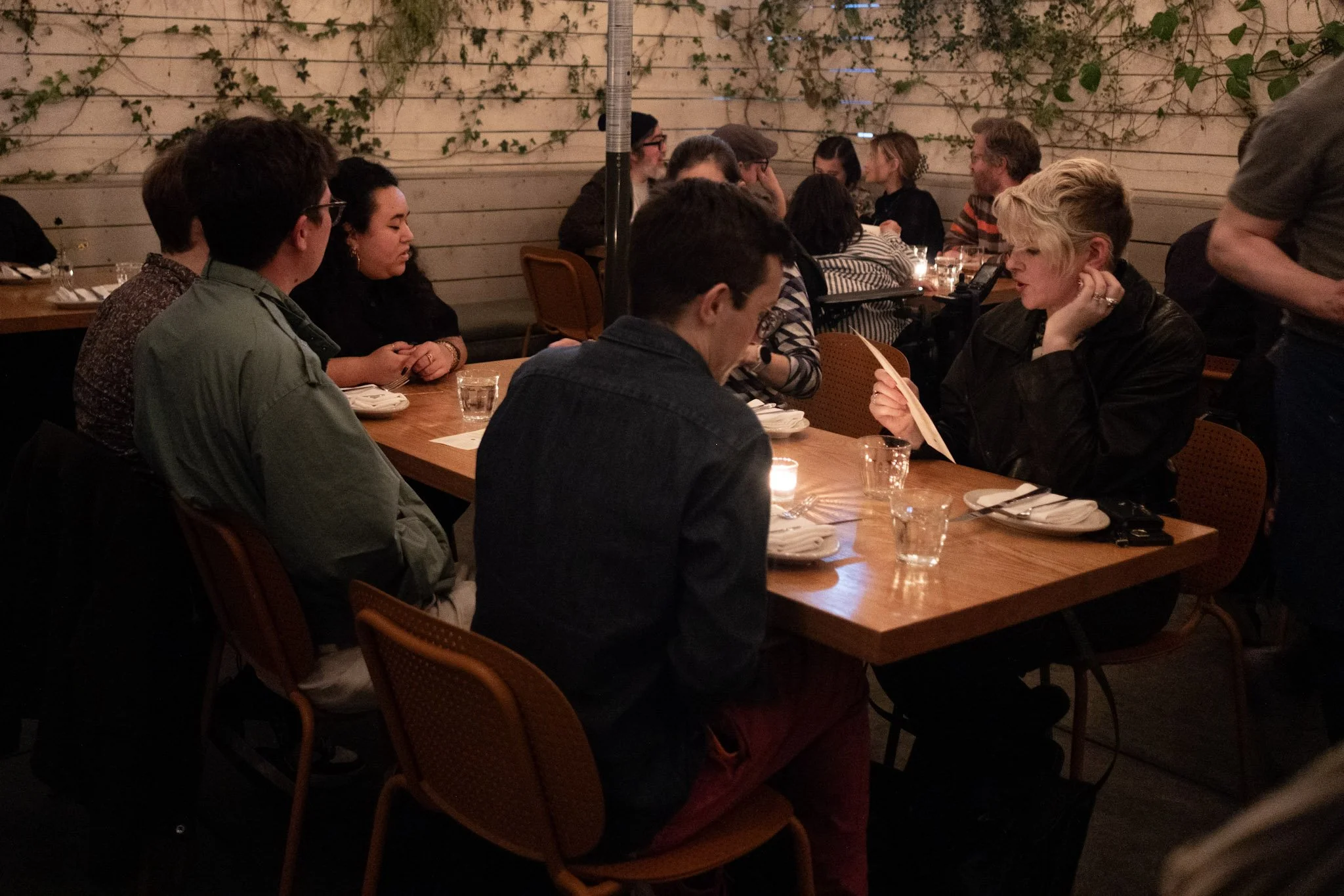  A cozy restaurant with people gathered around wooden tables. Dim lighting and climbing plants create an intimate atmosphere. Guests engage in conversation. 