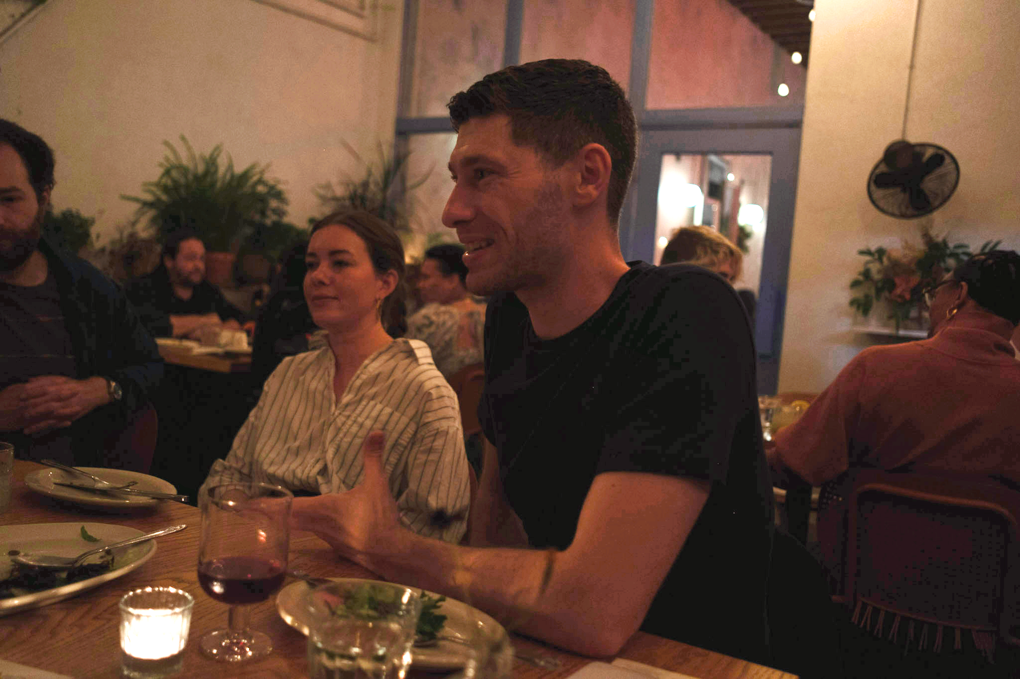  A group dinner in a cozy restaurant. A man gestures while talking, with a woman listening beside him.  