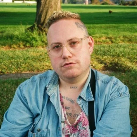 A Klass, a white queer/trans person dressed in light blue denim and a floral shirt, sits and stares into the camera with a grass field and tree behind them.