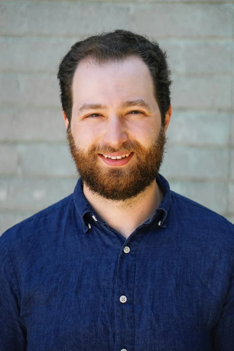 A headshot of a smiling white man with brown hair and a thick beard, wearing a blue button up shirt against a brick background.