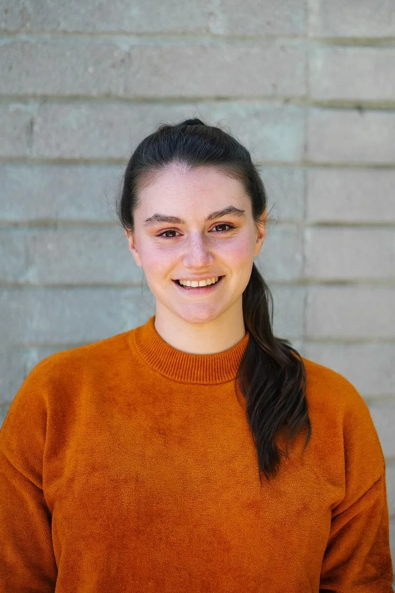 A headshot of a smiling white woman with brown hair in a ponytail, wearing an orange sweater.