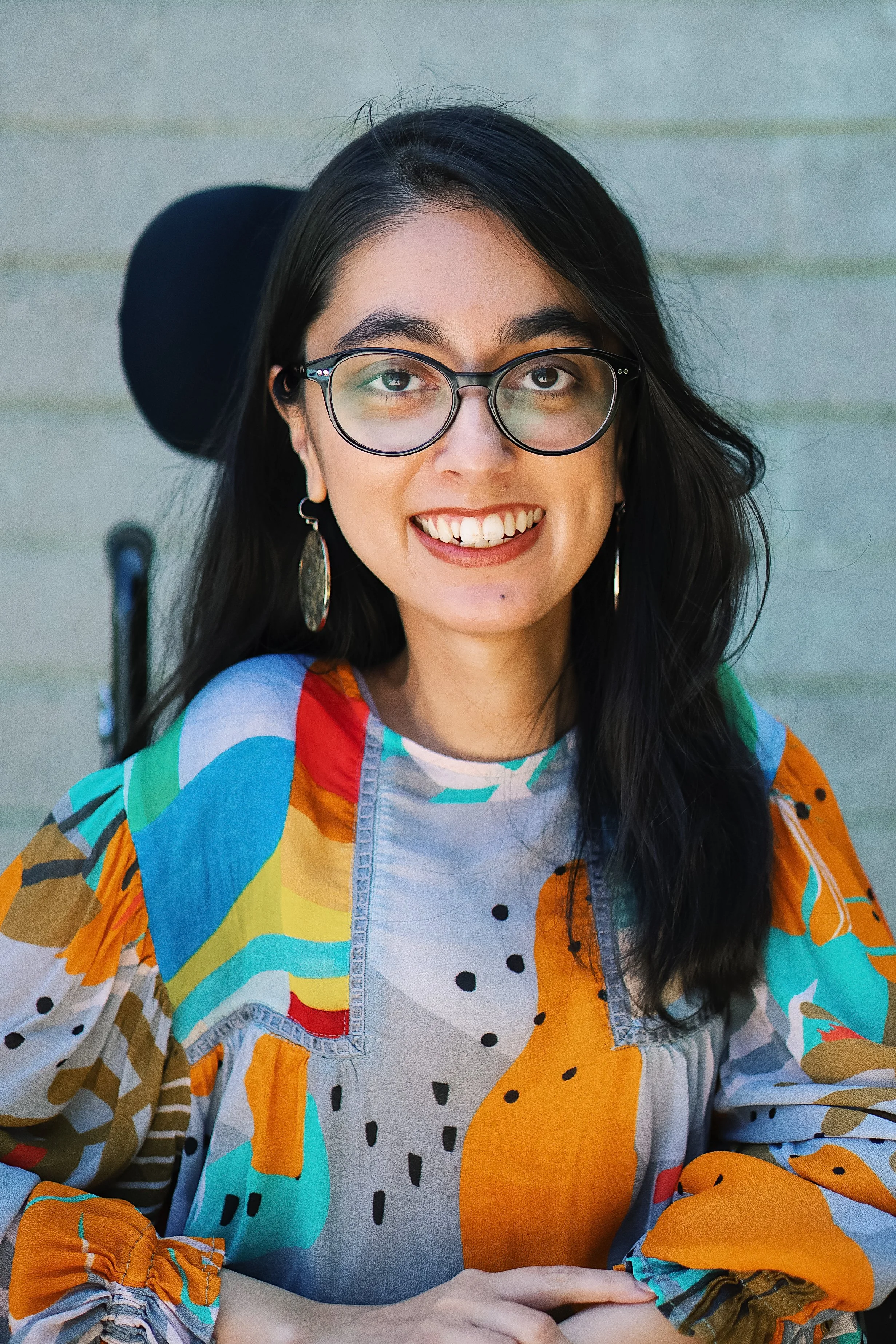 A South Asian woman with long black hair, black glasses, wearing a multi-colored blouse against a gray wall smiles at the camera.
