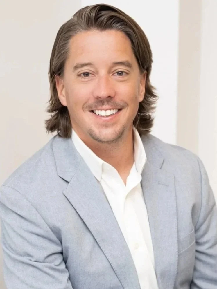 A white man with long brown hair, smiling at the camera, He's wearing a white collared shirt and a light blue blazer.