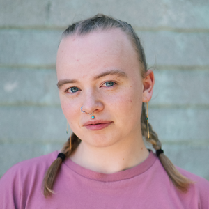 A headshot of a white person with blonde hair in braids and an upper lip and nose piercing. They wear a pink t-shirt.