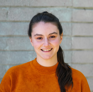 A headshot of a smiling white woman with brown hair in a ponytail, wearing an orange sweater.