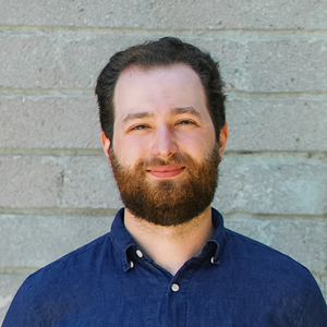 A headshot of a smiling white man with brown hair and a thick beard, wearing a blue button up shirt against a brick background.