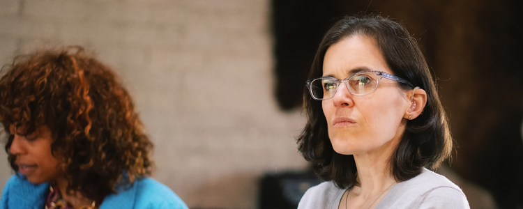 A photograph of two women, showing just their heads. The women on the left is out of focus and looks down and away from the camera. She is Black with curly brown hair and is wearing a blue jacket. The other woman is in focus and looks ahead at something in front of her. She has dark shoulder length hair and wears glasses and a hearing aid.