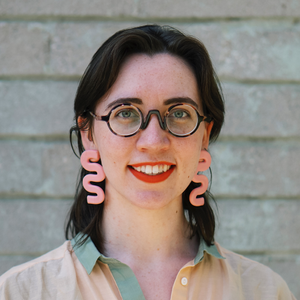 A headshot of a smiling white person with shoulder length brown hair. They wear glasses, pink squiggly earrings and a green and orange shirt.