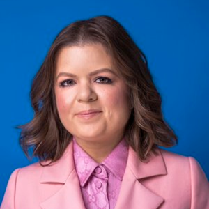 A headshot of a white woman with brown shoulder length hair, wearing a pink suit and shirt.