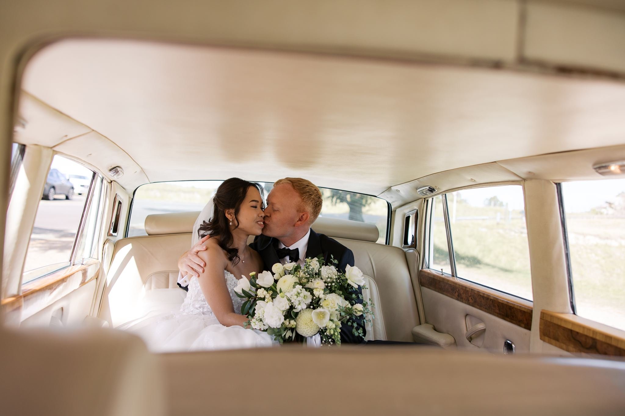 A bride and groom sharing a kiss inside a vintage car, with the bride holding a bouquet of white flowers.