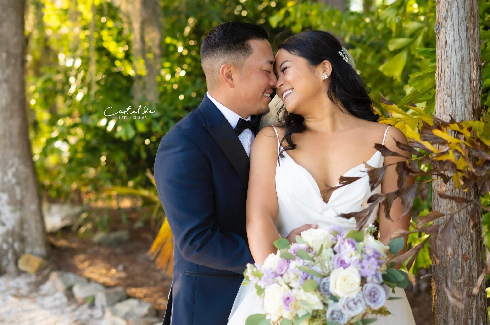 A newlywed couple sharing an intimate moment outdoors, with the groom in a dark tuxedo and the bride in a white dress holding a bouquet of white and purple flowers, surrounded by green trees and plants.