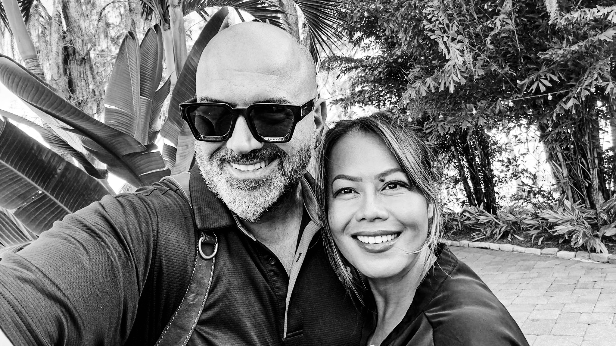 A smiling man and woman taking a selfie outdoors, with lush tropical plants in the background.