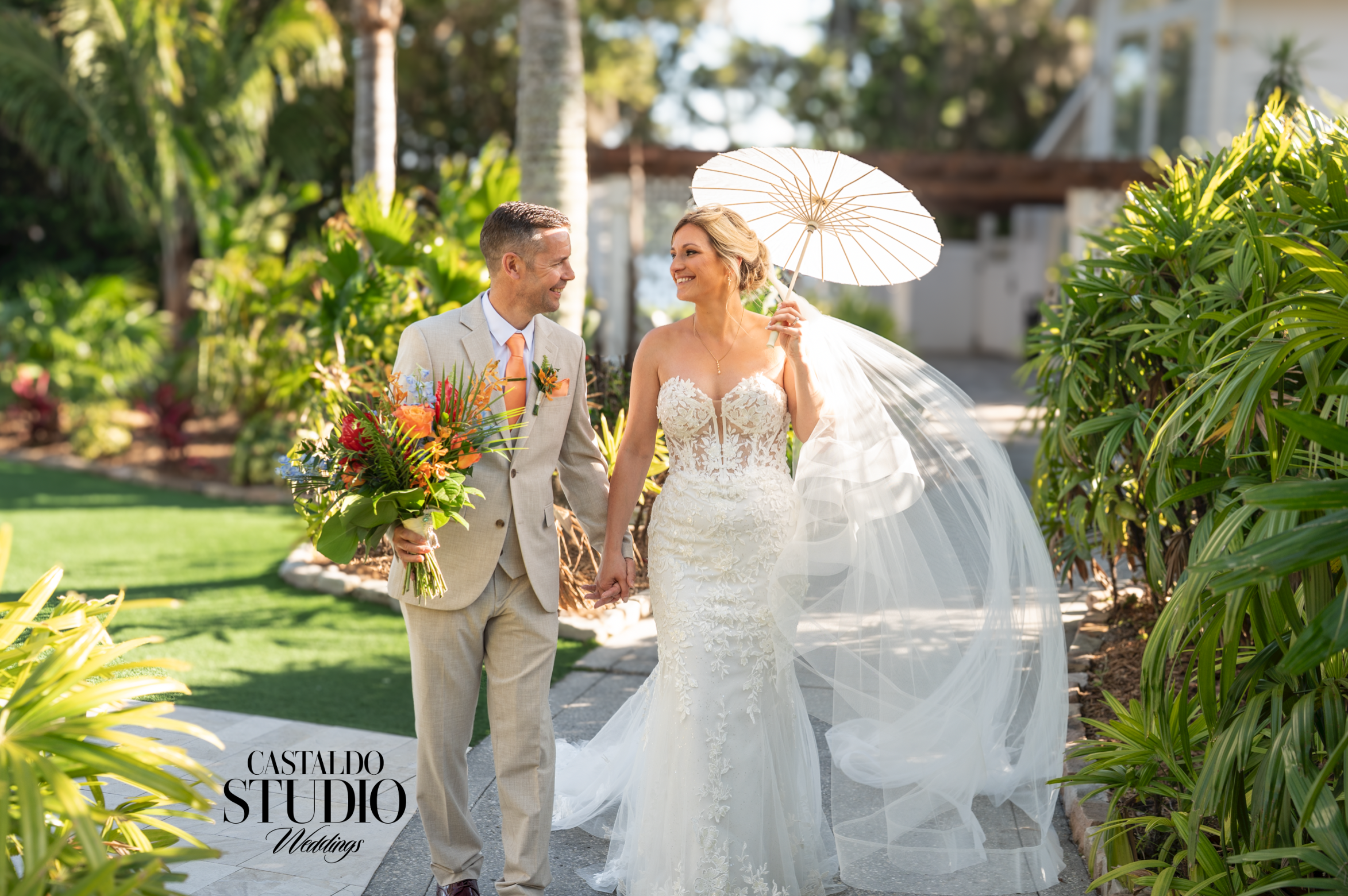 Bride in lace mermaid gown holding white parasol and groom in tan suit walking hand in hand through the tropical gardens at Paradise Cove Orlando, photographed by Castaldo Studio Weddings, Orlando destination wedding photographers.