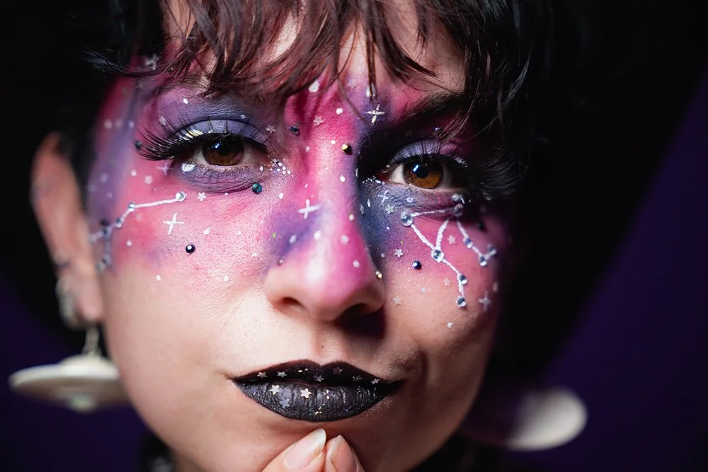 Close-up of a person with colorful galaxy-themed makeup and decorative facial embellishments, including star and constellation designs, black lipstick, and eye makeup, against a dark background.