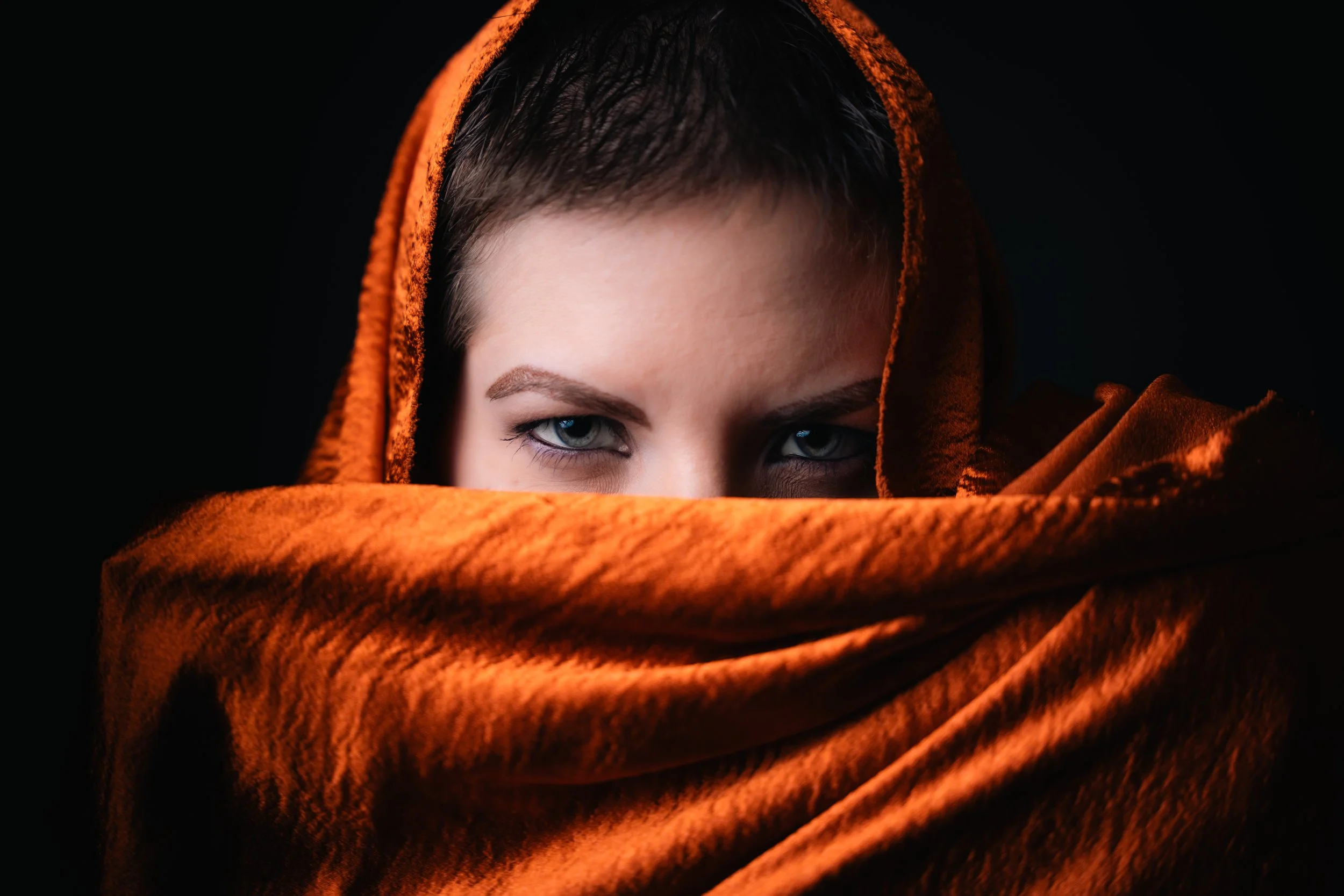 Close-up of a person's face with piercing blue eyes, wrapped in an orange fuzzy blanket with a dark background.