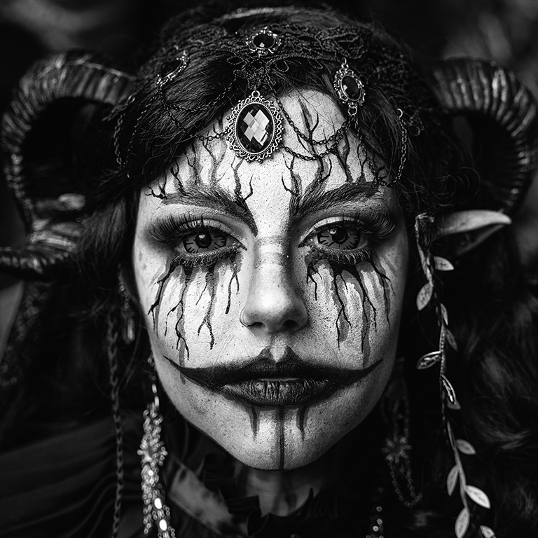 A person dressed as a fantasy character with elaborate makeup, horns, and jewelry, featuring dark lines and designs around the eyes and mouth in black and white.