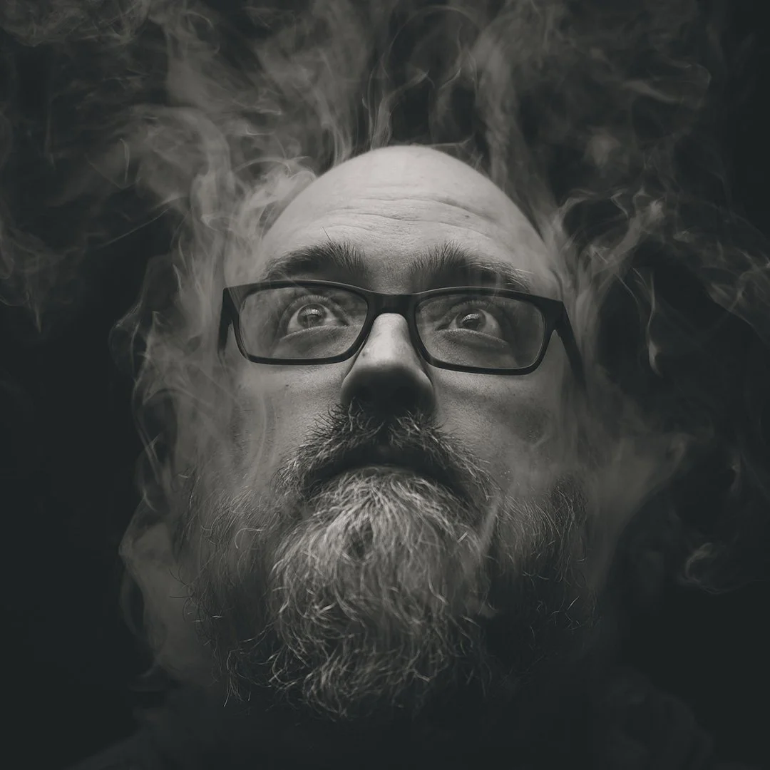 A black and white close-up photo of a man with glasses and a beard, looking upward with swirling smoke around his head.