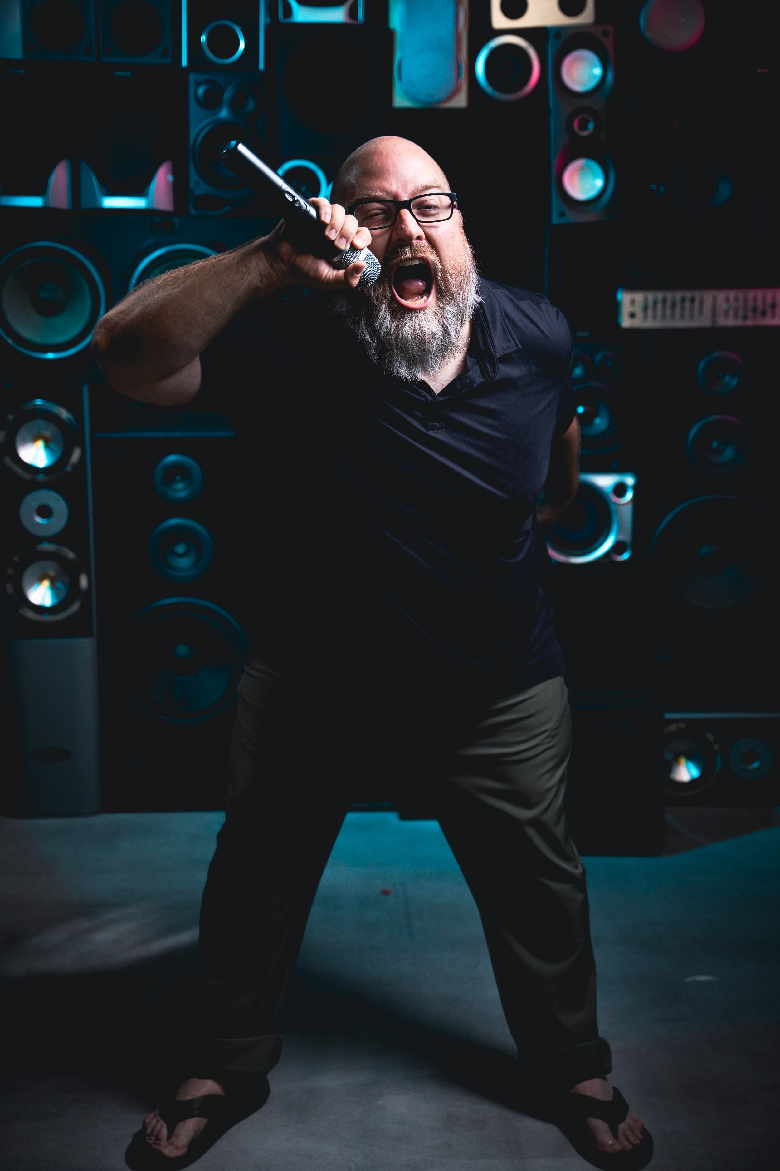 Bearded man wearing glasses, holding a microphone, shouting passionately, in front of a wall of large speakers, wearing black shirt and khaki pants.