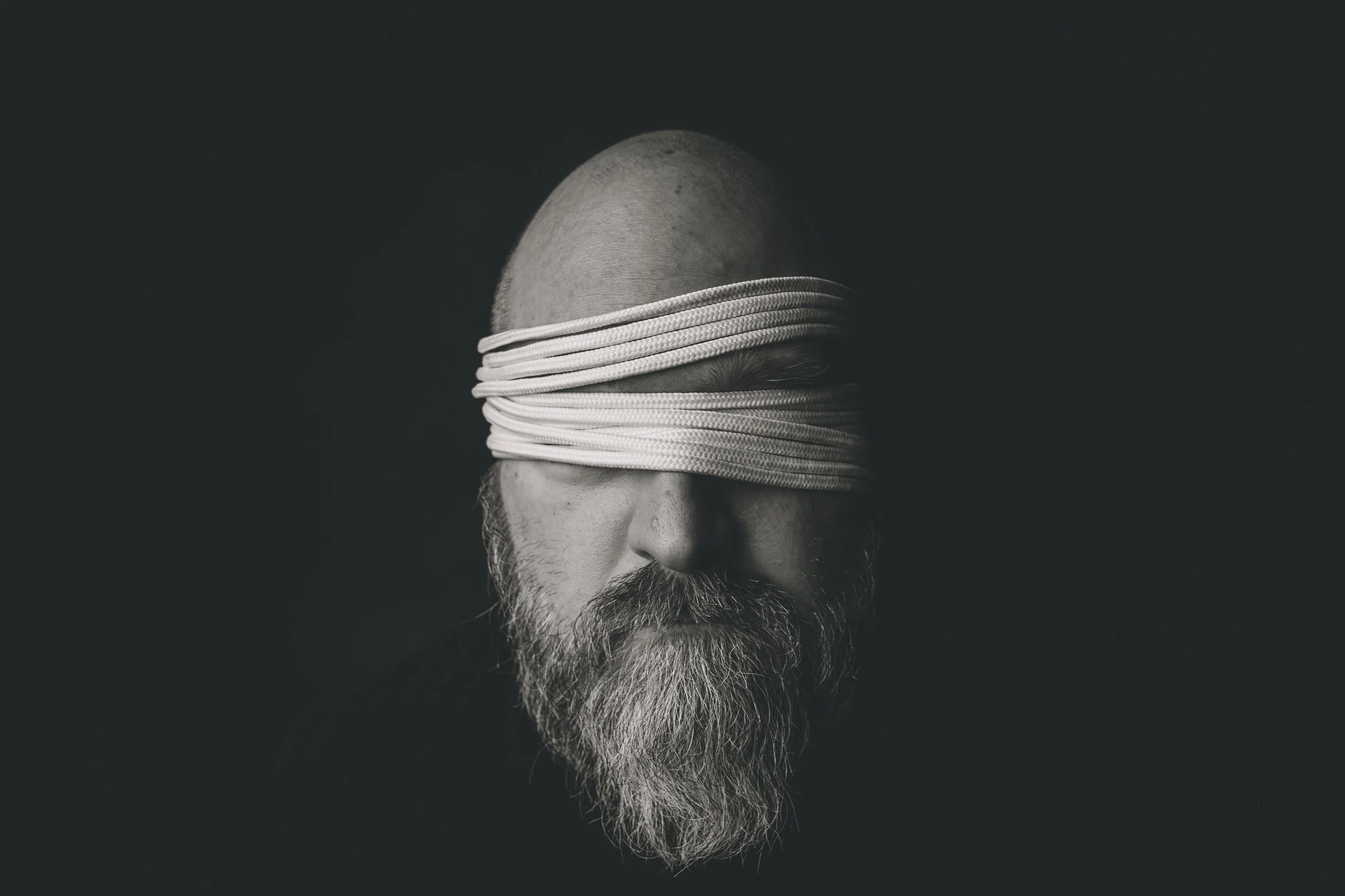 A black and white photo of a man with a bald head and a beard, whose eyes are covered with a stack of white ropes.