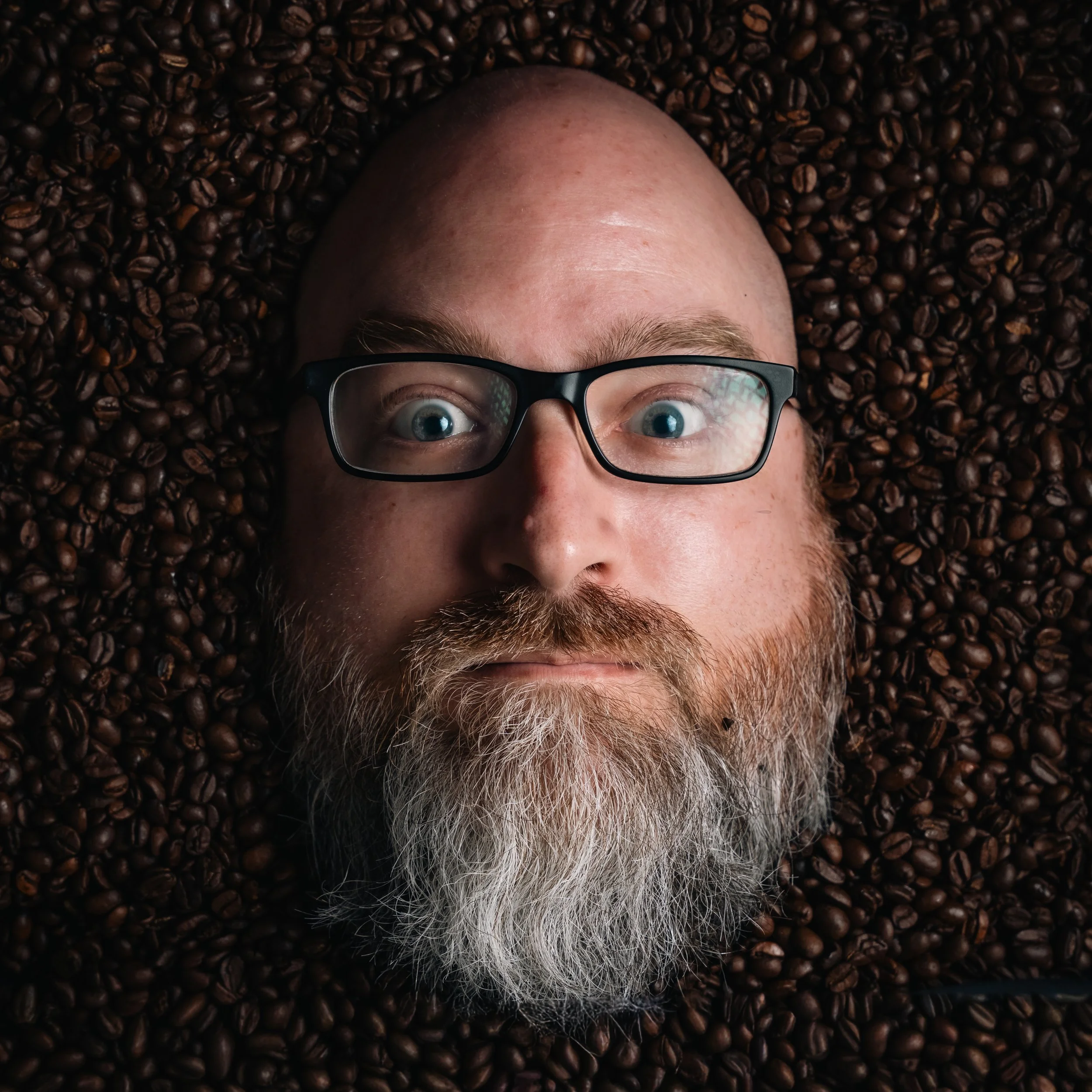 A person's face with glasses surrounded by dark roasted coffee beans.