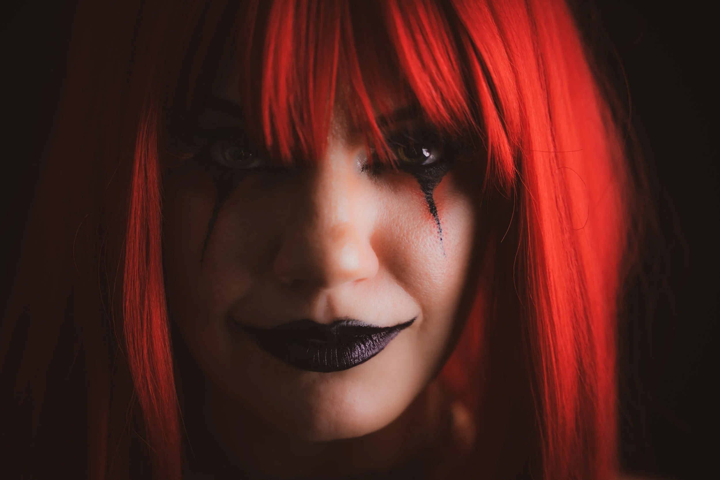 Close-up of a woman with bright red hair, dark makeup, black lipstick, and black tear-like streaks under her eyes.