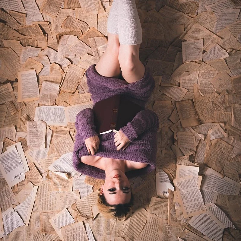 A woman lying down on the floor surrounded by scattered open books, wearing a purple sweater and white knee-high socks.