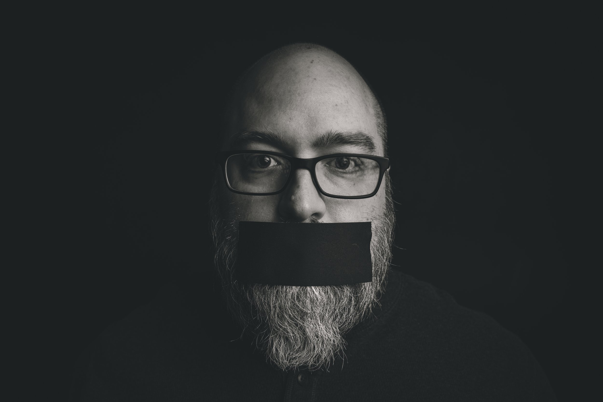 Black and white portrait of a bald man with glasses and a beard, with his mouth covered by a black strip.
