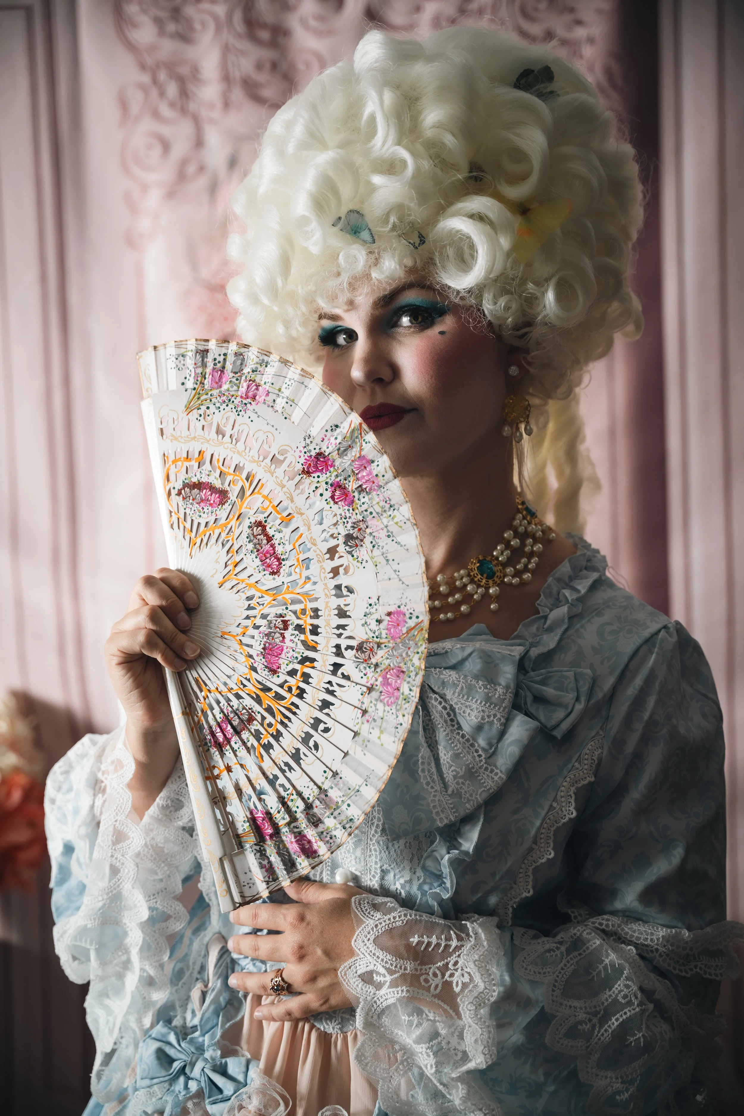 A woman dressed in 18th-century European costume with elaborate white wig holding a decorated fan in a vintage-style room.