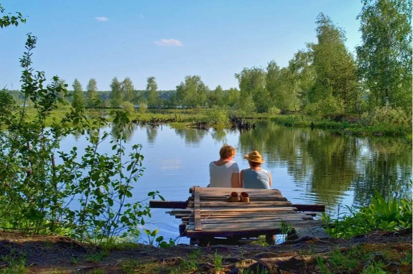 Two people sitting on a wooden dock by a tranquil lake surrounded by lush green trees.