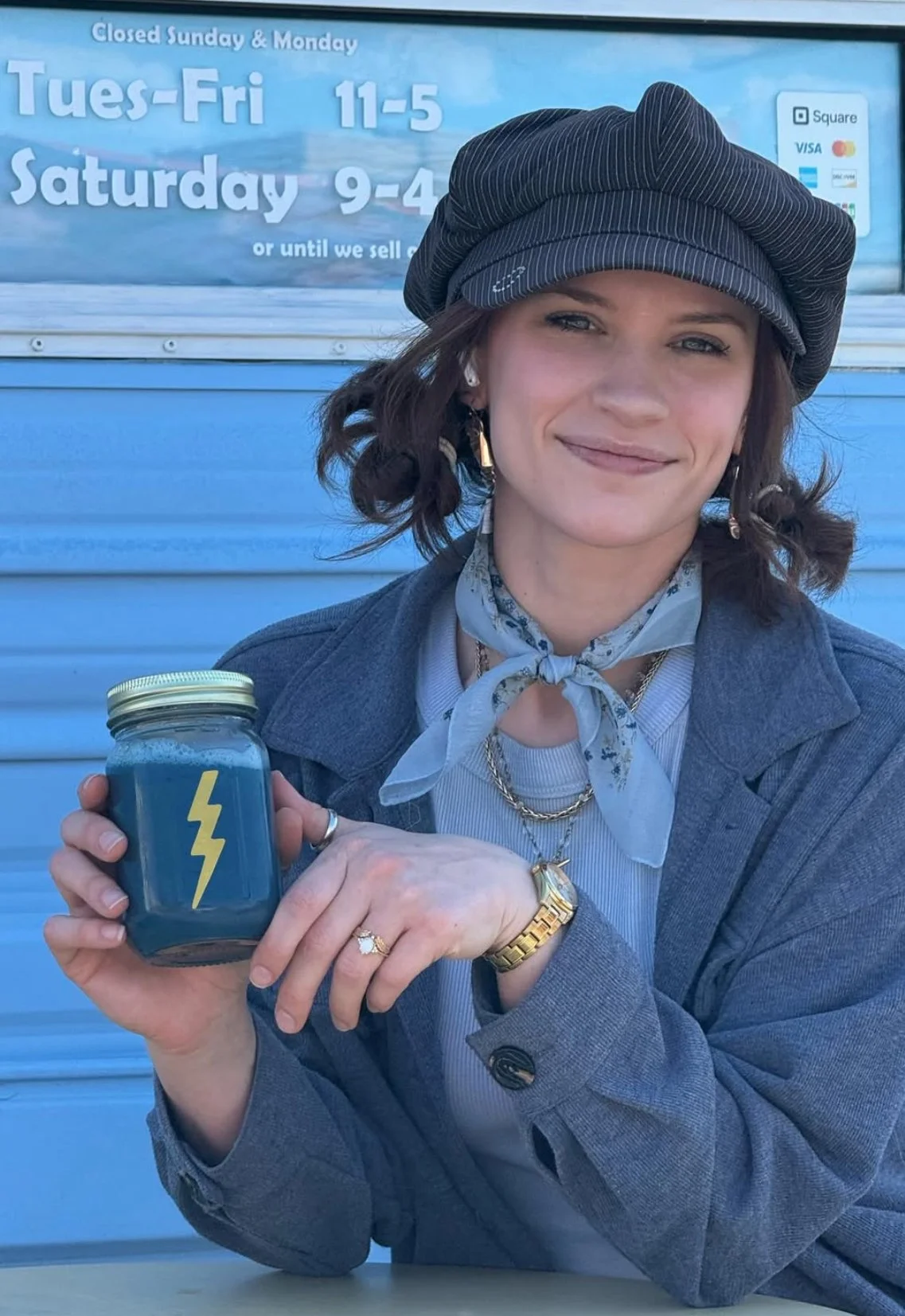 A woman with dark wavy hair, wearing a black beret, gray blazer, striped shirt, gold jewelry, and a light scarf, holds a jar with a yellow lightning bolt symbol, sitting outside with a blue wall and a sign displaying business hours in the background.