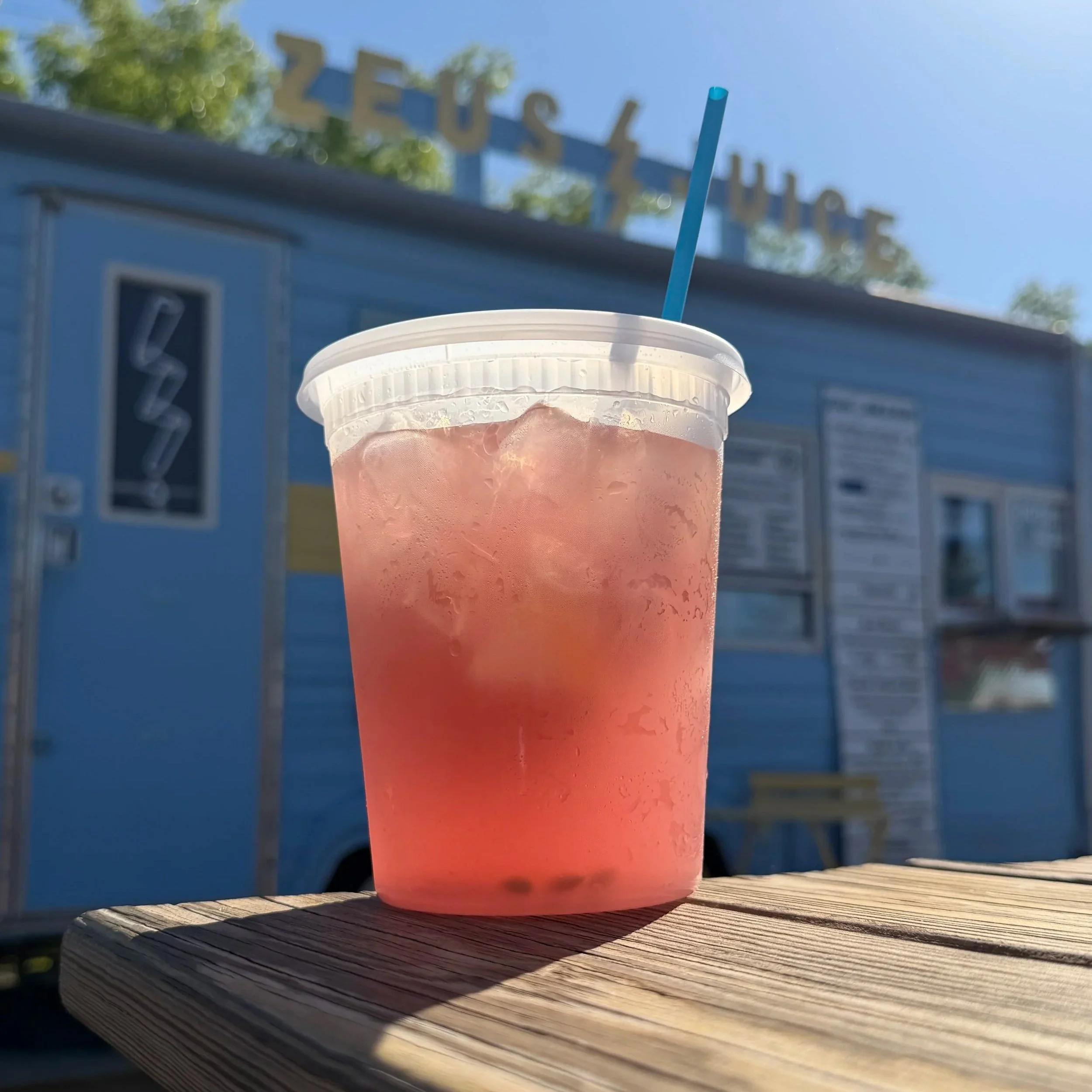 Freshly Squeezed lemonade. Hibiscus Flavored. A clear plastic cup with a white lid and blue straw, filled with pink iced beverage, sitting on a wooden surface outdoors with a blue food truck in the background.
