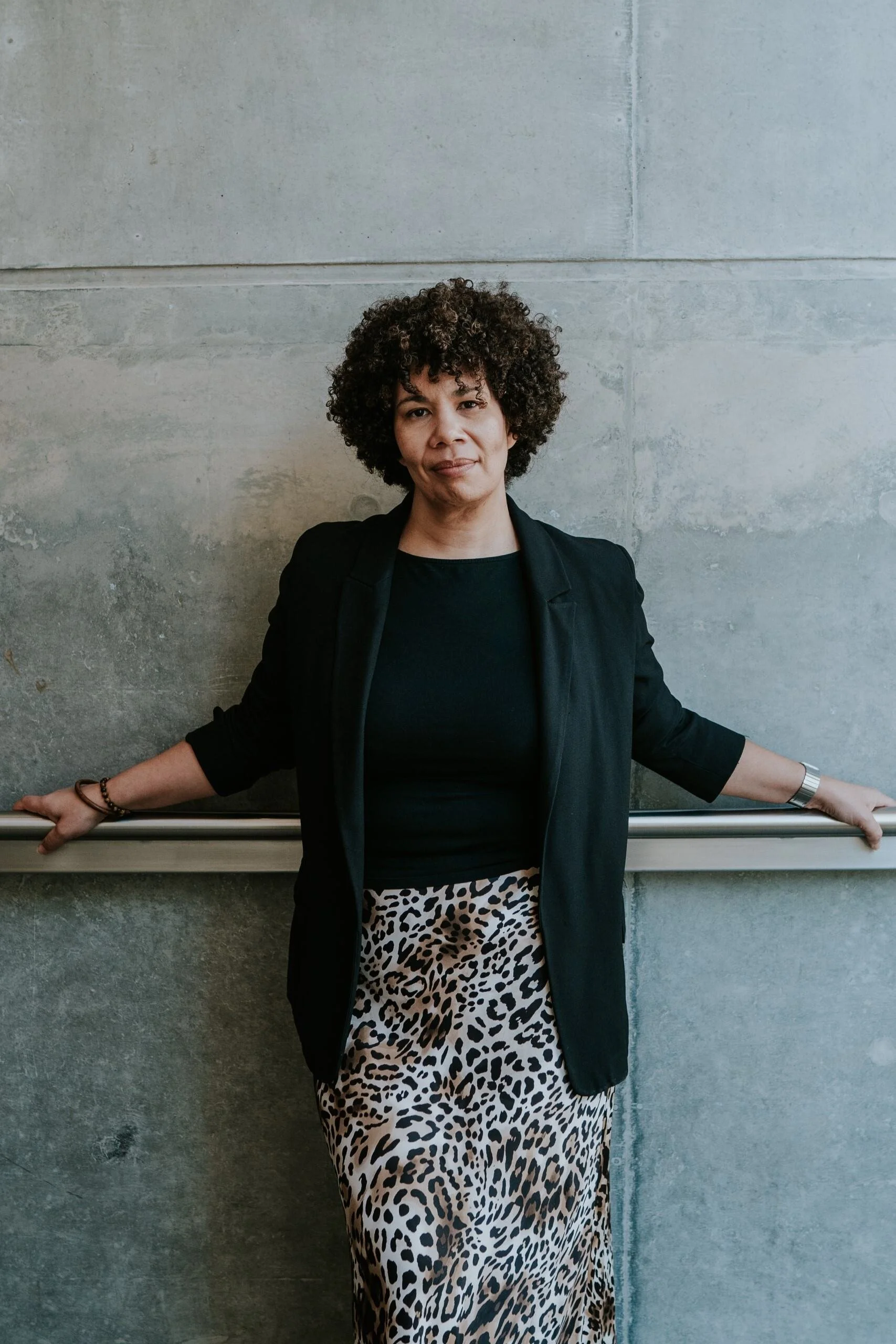 A woman with curly hair wearing a black blazer, black shirt, and leopard print skirt poses with arms extended, holding onto a railing against a concrete wall.
