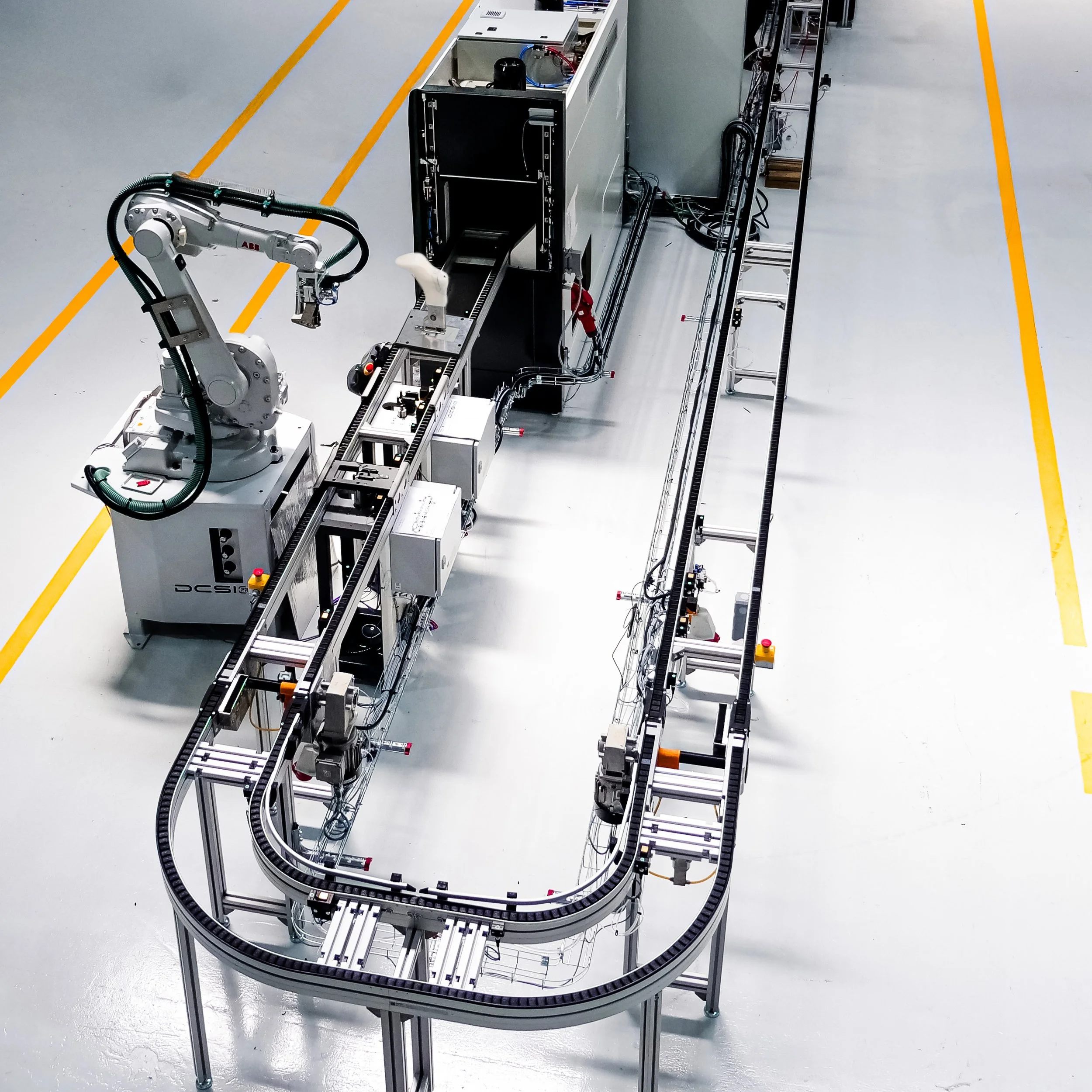 Robô industrial com braço articulado operando em uma linha de montagem de componentes eletrônicos ou dispositivos em um ambiente de fábrica limpa com piso de cor cinza claro e linhas amarelas de demarcação.