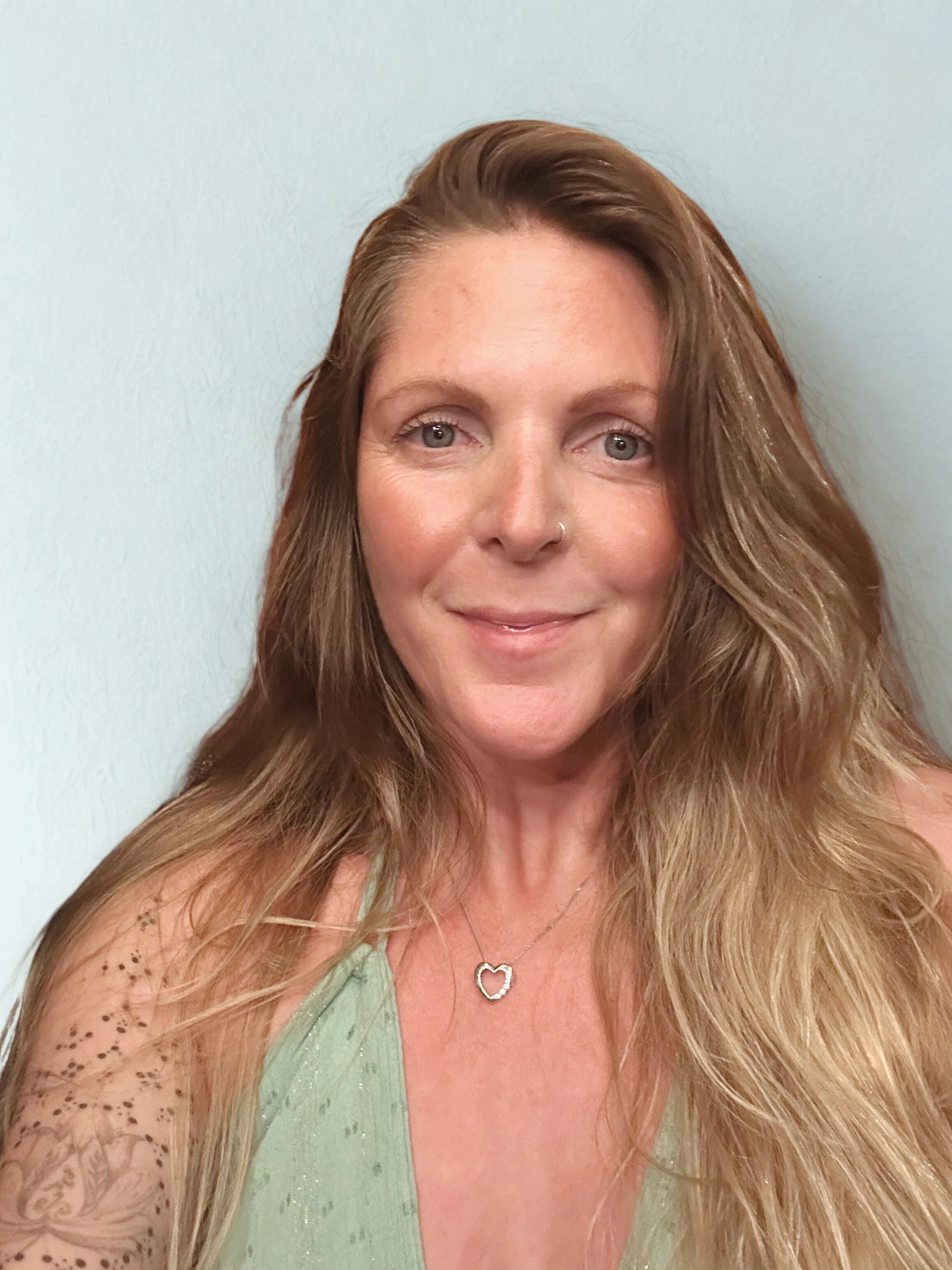 A white woman with long, wavy, reddish-brown hair, wearing a silver heart-shaped necklace and a green top, smiling against a light-colored background.