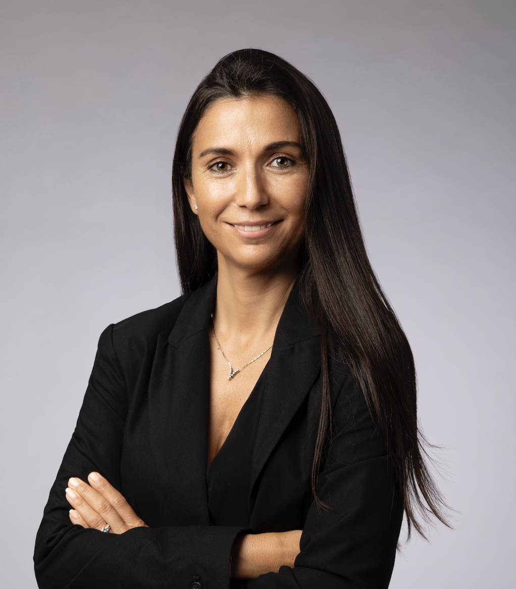 Professional woman with long dark hair and a black blazer, smiling and arms crossed, against a plain light gray background.